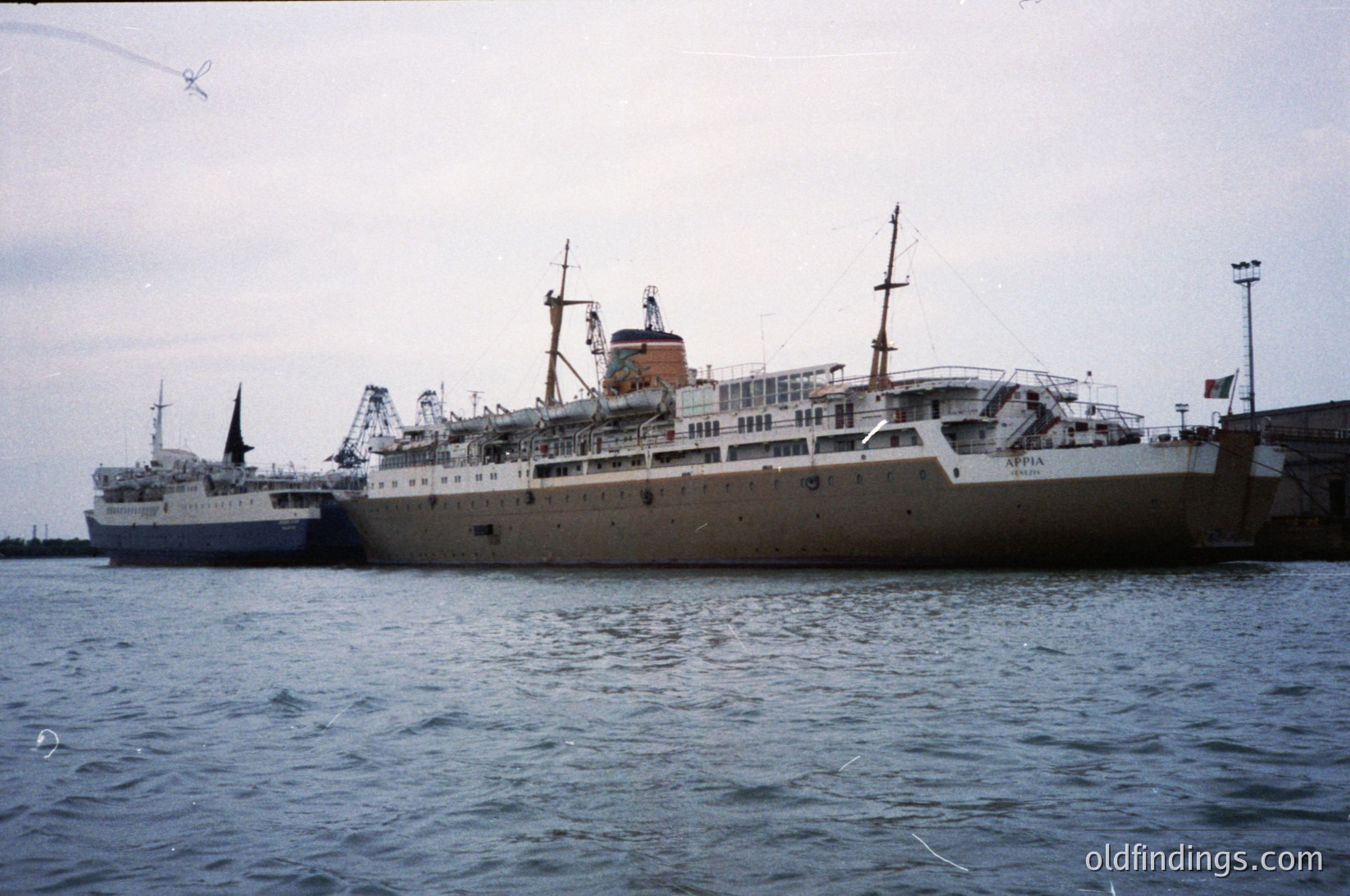 Mid-20th century passenger ship docked at port, likely –. White hull with red-orange funnel, multiple decks, and lifeboats visible. Industrial port infrastructure with cranes and concrete piers in background. Potential maritime history or shipping archive reference.