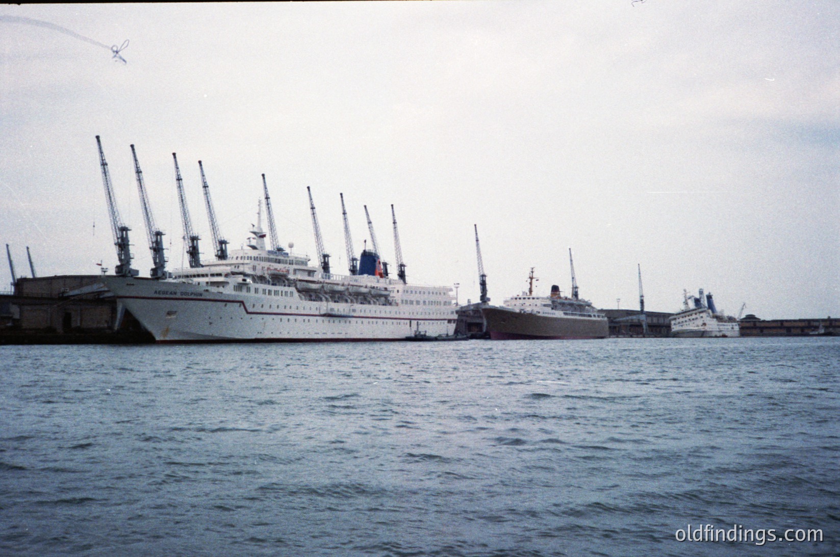 Mid-20th century ocean liner docked at port, likely 1950s–1960s. Multi-deck vessel with tall masts and white hull, flanked by smaller ships. Industrial port infrastructure visible in background. Ideal for maritime history, vintage travel, or maritime design research.