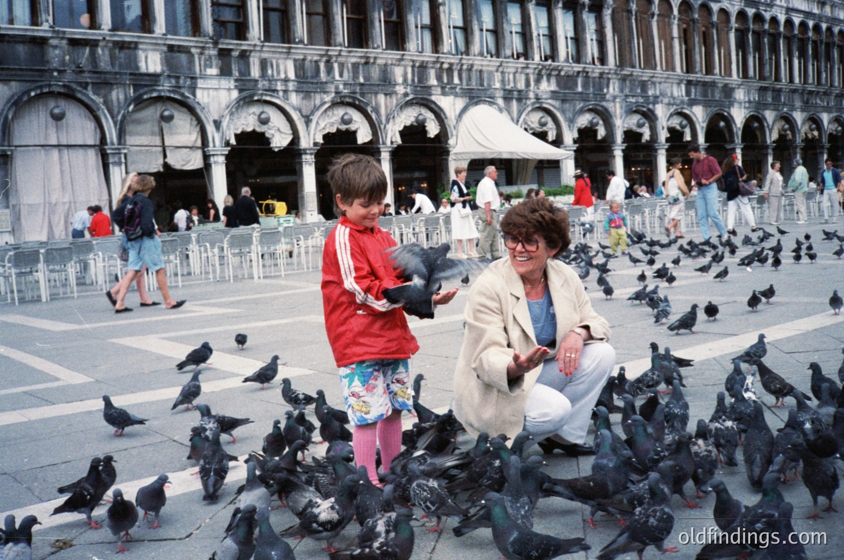 A woman and child feed pigeons in St. Mark’s Square (), Venice, Italy. Architectural details include arcaded loggias and Renaissance-style facades. Mid-20th century attire suggests or . Crowds and pigeons highlight iconic urban life.