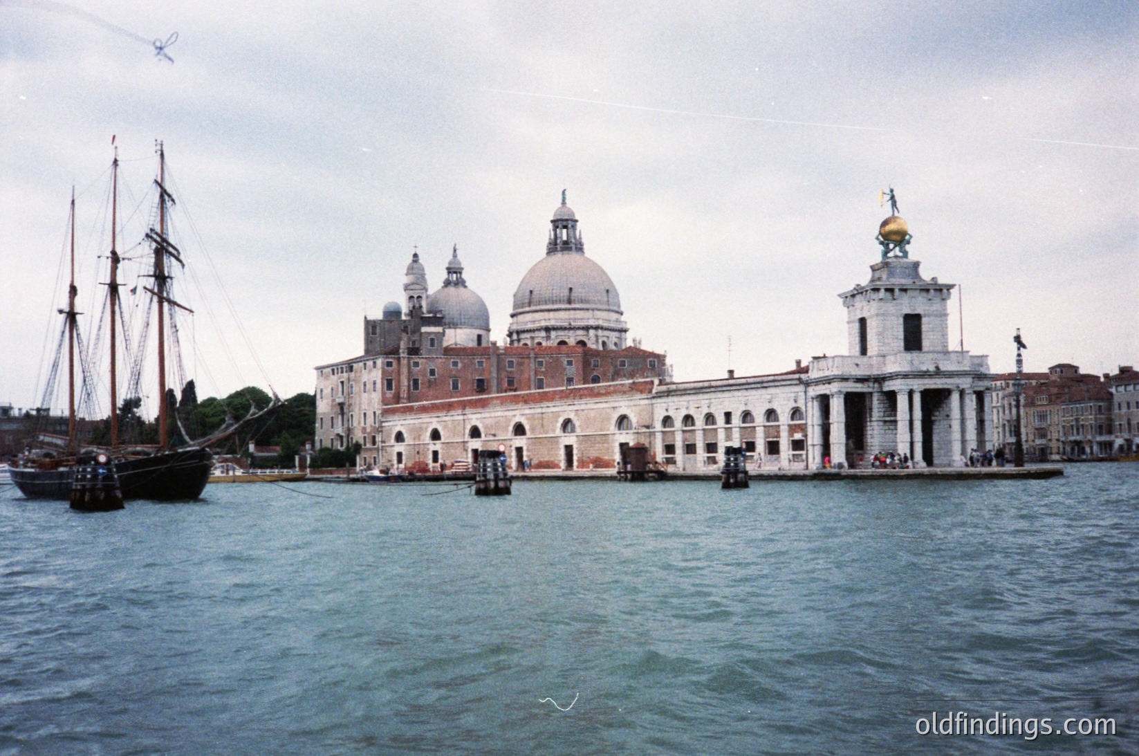 Historic Venetian waterfront featuring the **San Giorgio Maggiore** church with its iconic domes and classical façade. A traditional wooden sailing ship docked alongside the island’s stone pier. Overcast sky enhances the timeless atmosphere. Likely late 20th century.
