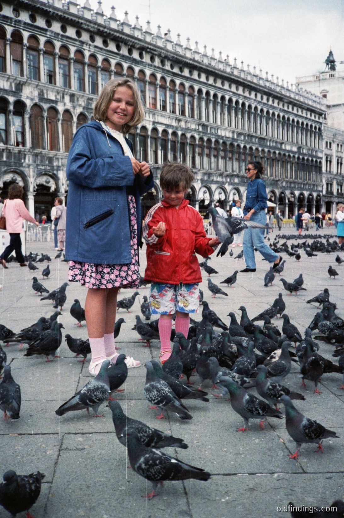 Two children feed pigeons in St. Mark’s Square (), Venice, Italy. The adult in blue jacket and floral skirt stands beside a child in red jacket and patterned pants, surrounded by a flock of pigeons. Renaissance-era Venetian Gothic architecture with arched windows and ornate stonework forms the backdrop. Likely late 20th century (–1980s).