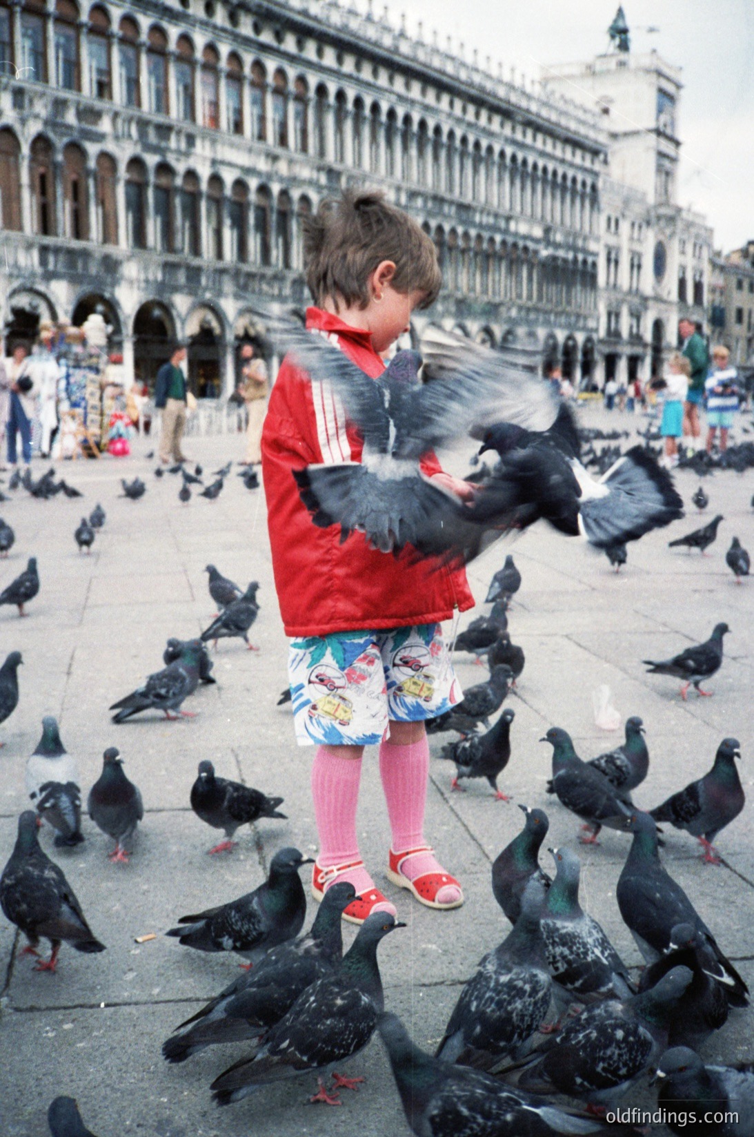 A child in a red jacket and floral shorts feeds pigeons in St. Mark’s Square, Venice, Italy. The historic Venetian Gothic architecture of the Basilica di San Marco and Doge’s Palace frames the scene. Likely late 20th century (1970s–1990s).