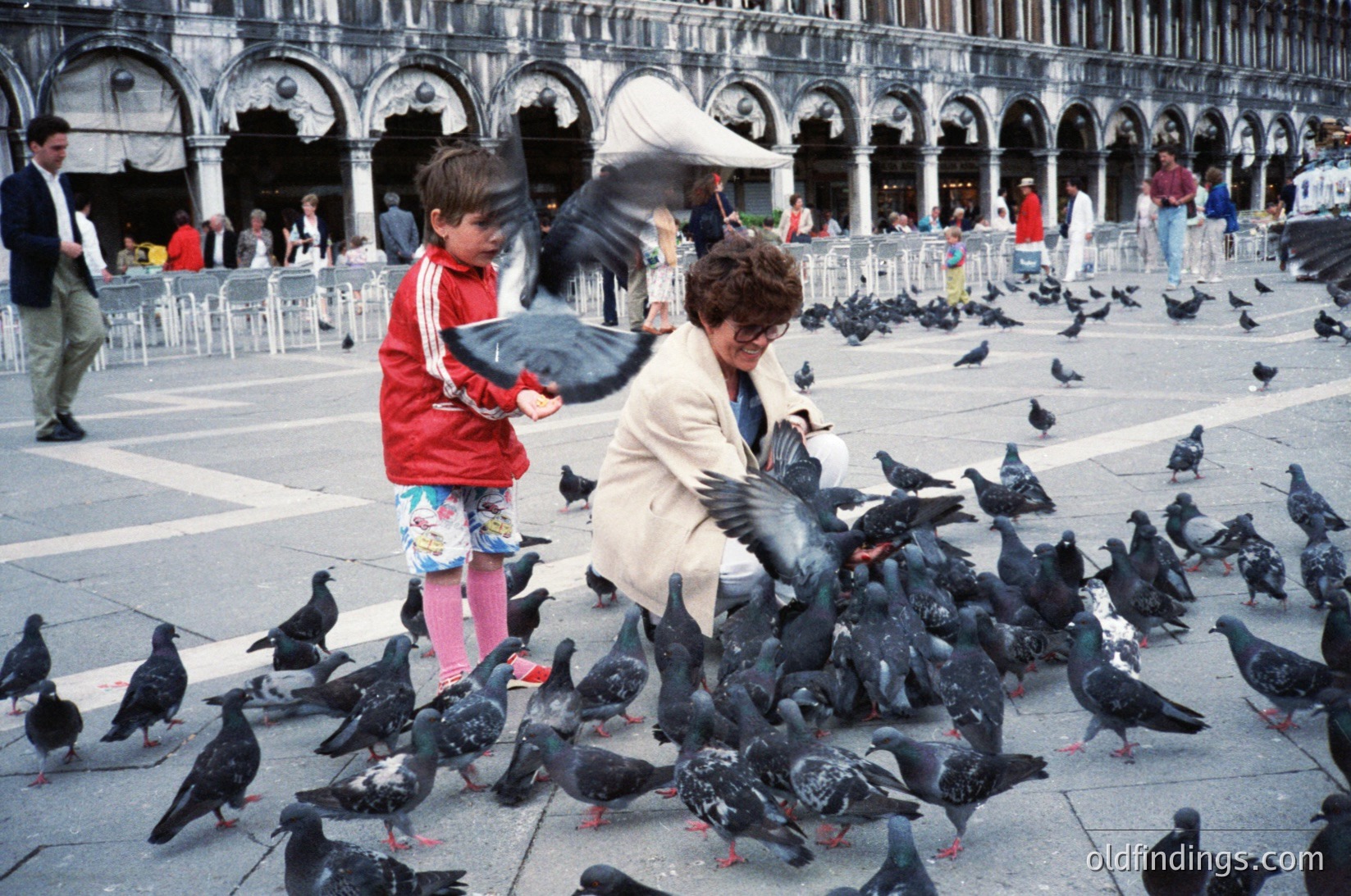 A woman and child feeding pigeons in St. Mark’s Square (), Venice, Italy. The 1970s-era scene captures iconic Venetian architecture with arcaded loggias and a crowd of black birds. Distinctive striped sweater and wide-brimmed hat add period detail.