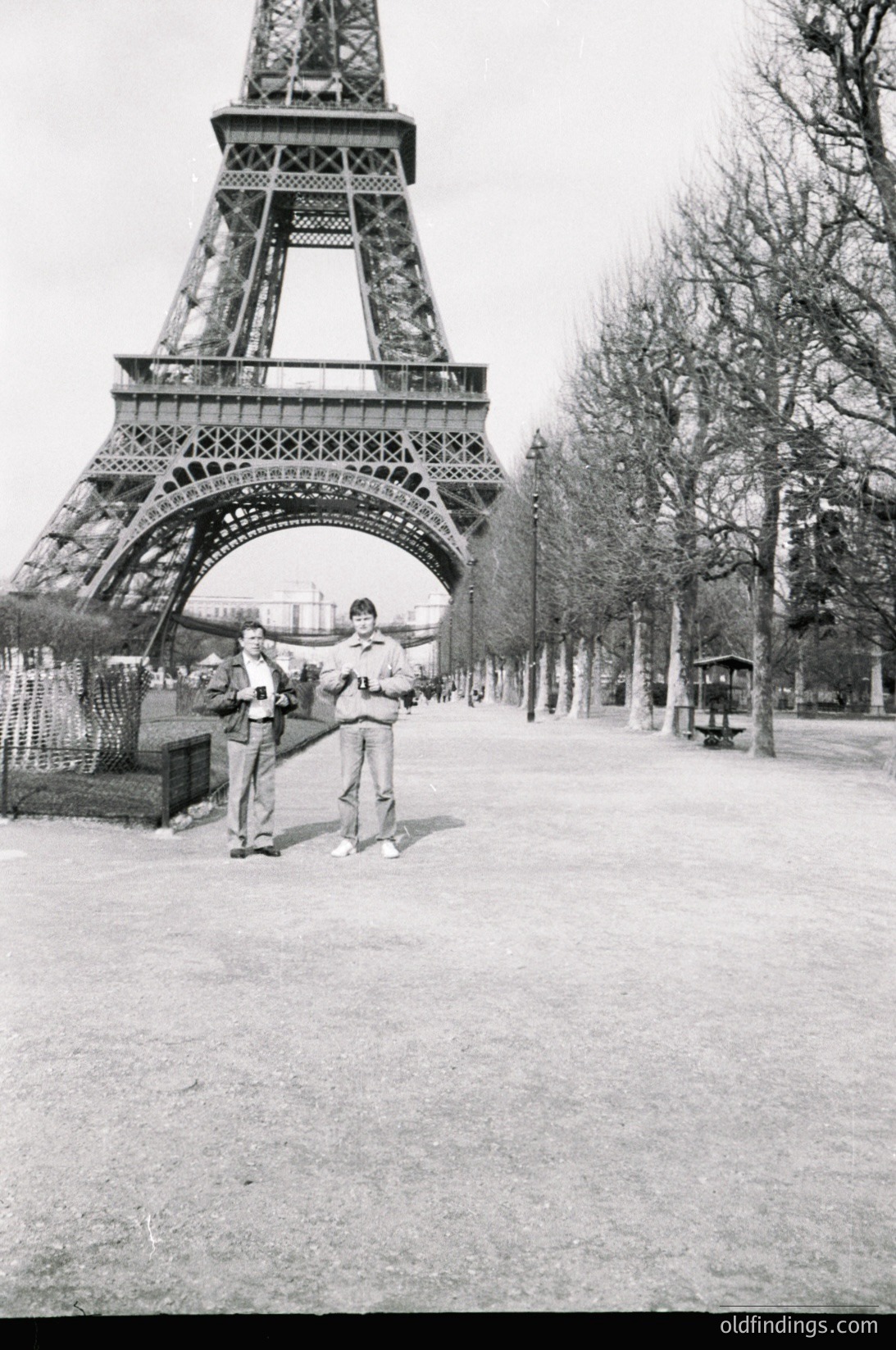 Two men in 1970s-style clothing pose near the Eiffel Tower’s base in Paris, likely Champs de Mars. One wears a light jacket and bell-bottoms; the other, a dark jacket and tie. Bare trees and a vintage tram shelter frame the scene.