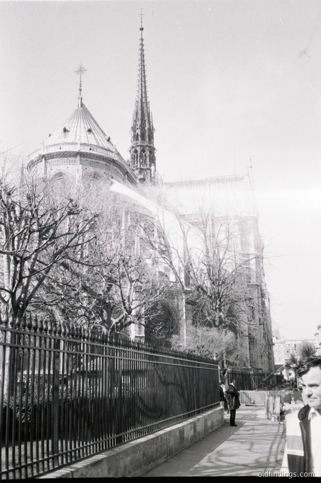 Gothic-style cathedral with twin spires and domed central tower, partially obscured by mist. Iron fence and bare trees frame the scene. Mid-20th century urban setting, likely European.