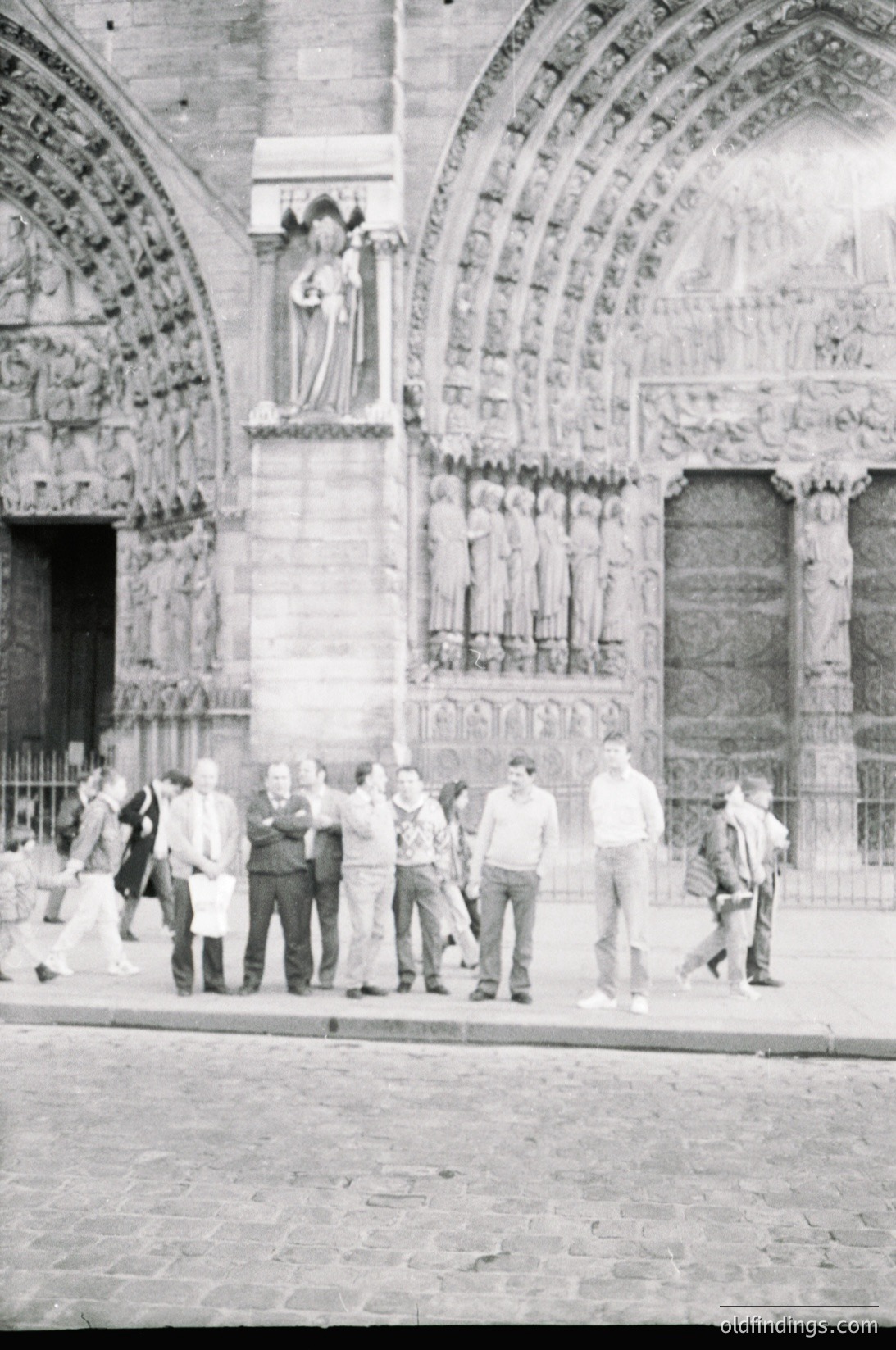 Romanesque-style cathedral façade featuring intricate stone carvings, arched doorways, and a central relief of a robed figure. Mid-20th century group of casually dressed individuals posing in front of the entrance.
