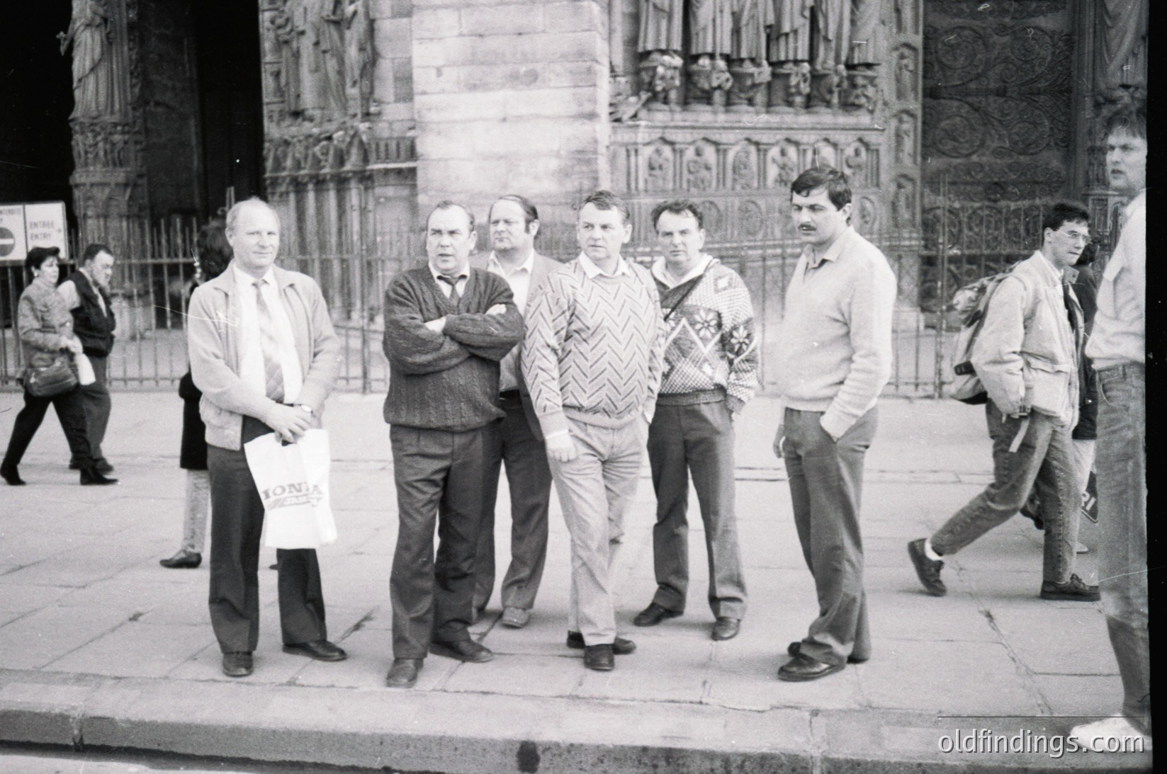 Group of six men posing in front of Gothic-style cathedral façade, likely 1970s–1980s. Distinctive herringbone sweater and rolled-collar shirts suggest European mid-century fashion. One man holds a sign with partial text "LOND...". Crowd and architectural details hint at European city center.