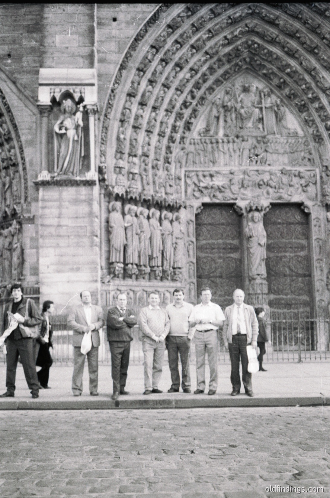 Gothic cathedral façade featuring intricate stone carvings, statues, and pointed arches. Group of men in 1960s-era clothing posing in front of ornate entrance. Cobblestone plaza and wrought-iron gates visible.