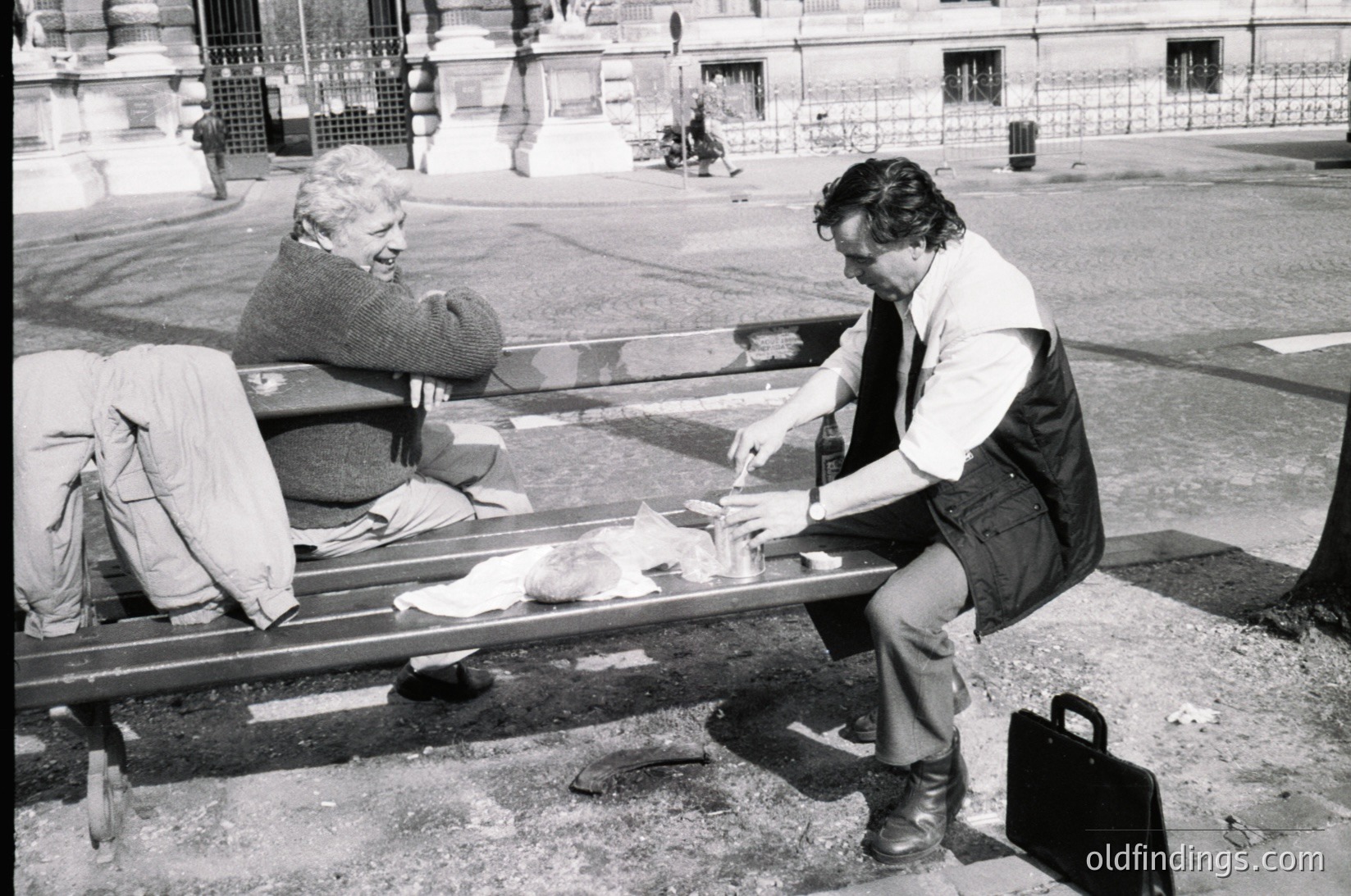 Two men share a meal on a park bench in an urban setting, likely mid-20th century. The seated man eats while the standing man holds a plate. Surrounding architecture includes wrought-iron fencing and stone walls. Casual, candid street life captured.