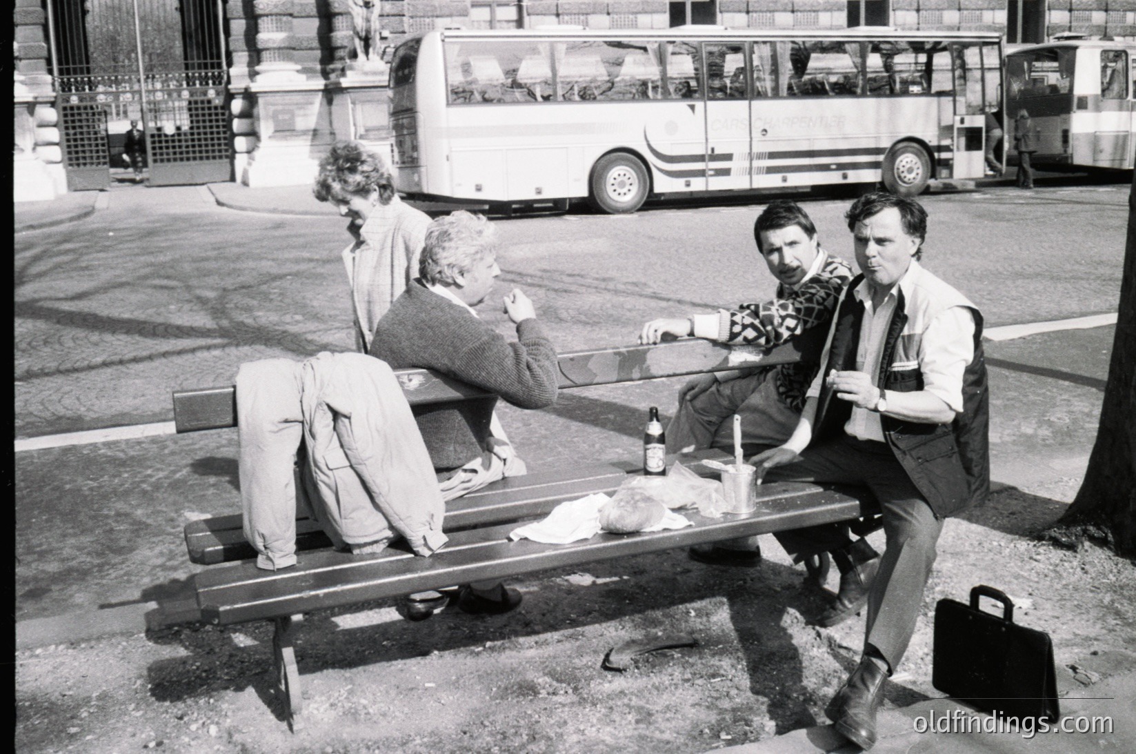Three men relax on a wooden bench in an urban setting, 1970s. One holds a bottle, another a briefcase; vintage bus and classic streetwear visible. Captures post-war European social dynamics.