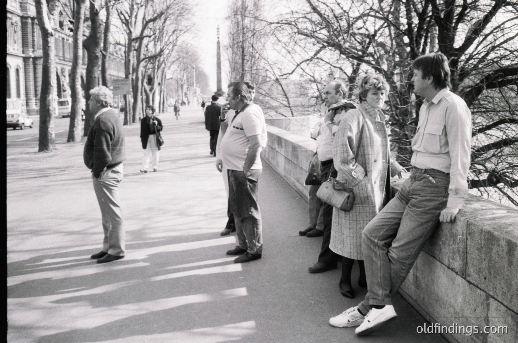 Group of five adults in 1970s streetwear—men in button-downs, women in layered coats—standing on a wide urban promenade lined with bare trees. Concrete barriers and distant pedestrians suggest a city park or boulevard. Classic black-and-white composition evokes mid-century European street life.