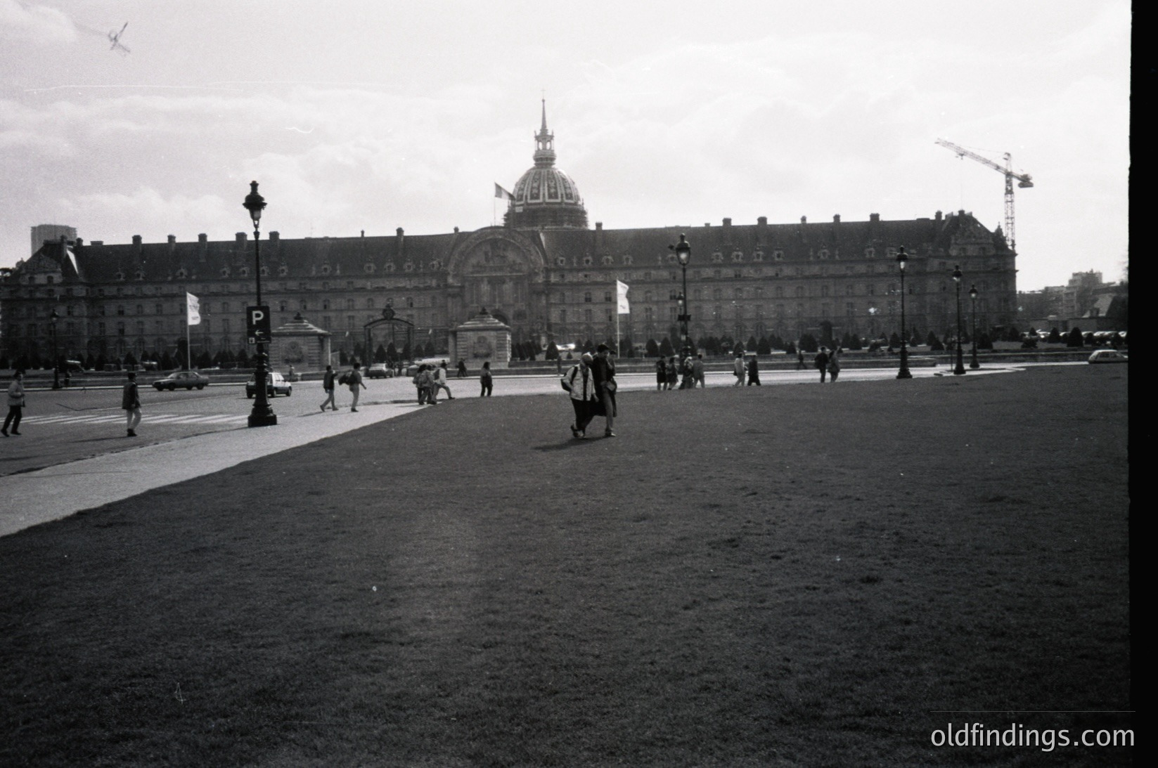 Neoclassical palace with domed roof and symmetrical façade, likely the Hôtel des Invalides in Paris. Crowded plaza with pedestrians, vintage cars, and lampposts. Construction crane visible at right. Black-and-white, mid-20th century (1950s–1960s). ôtelDesInvalides