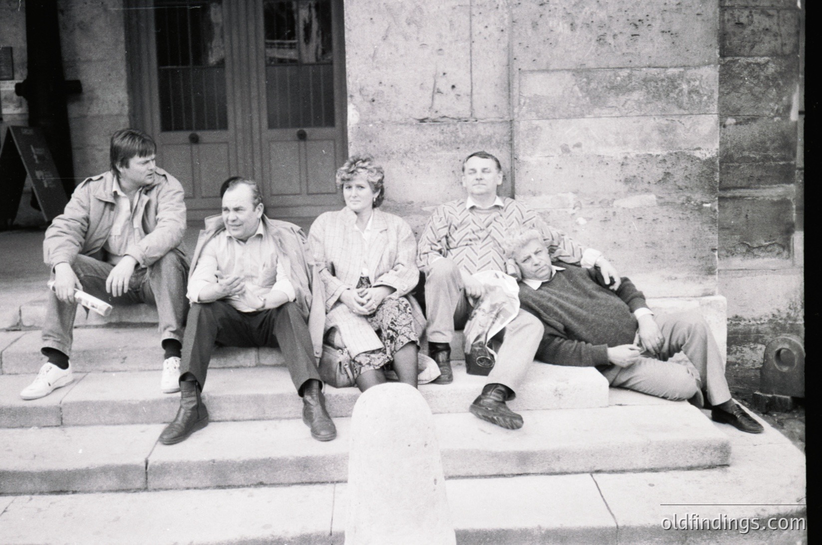 Four individuals pose casually on stone steps outside a stone-framed building, likely mid-20th century (1960s–1970s). Clothing includes button-ups, patterned shirts, and trousers—men’s fashion of the era. The woman wears a headband and a patterned blouse. Architectural details suggest institutional or public setting.