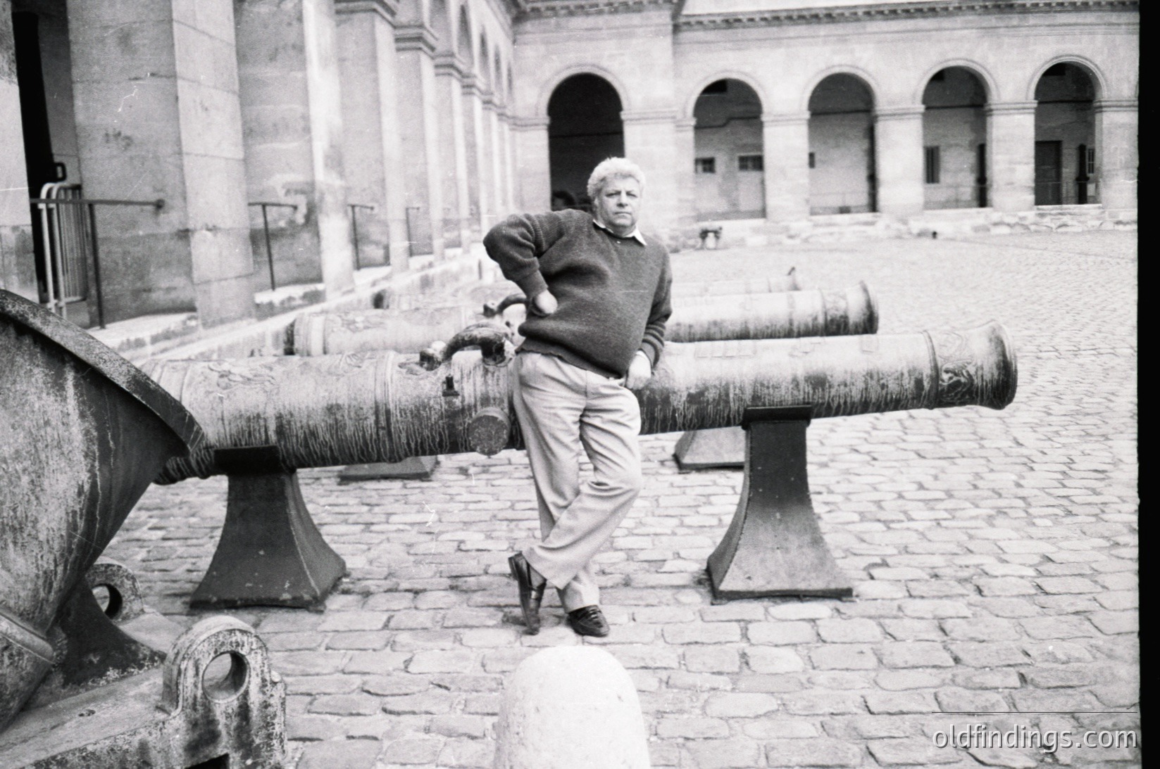 Man poses confidently atop 19th-century cannon in cobblestone courtyard, leaning on barrel with one hand. Architectural backdrop features arched colonnades and weathered stone. Likely European military heritage site, mid-20th century.