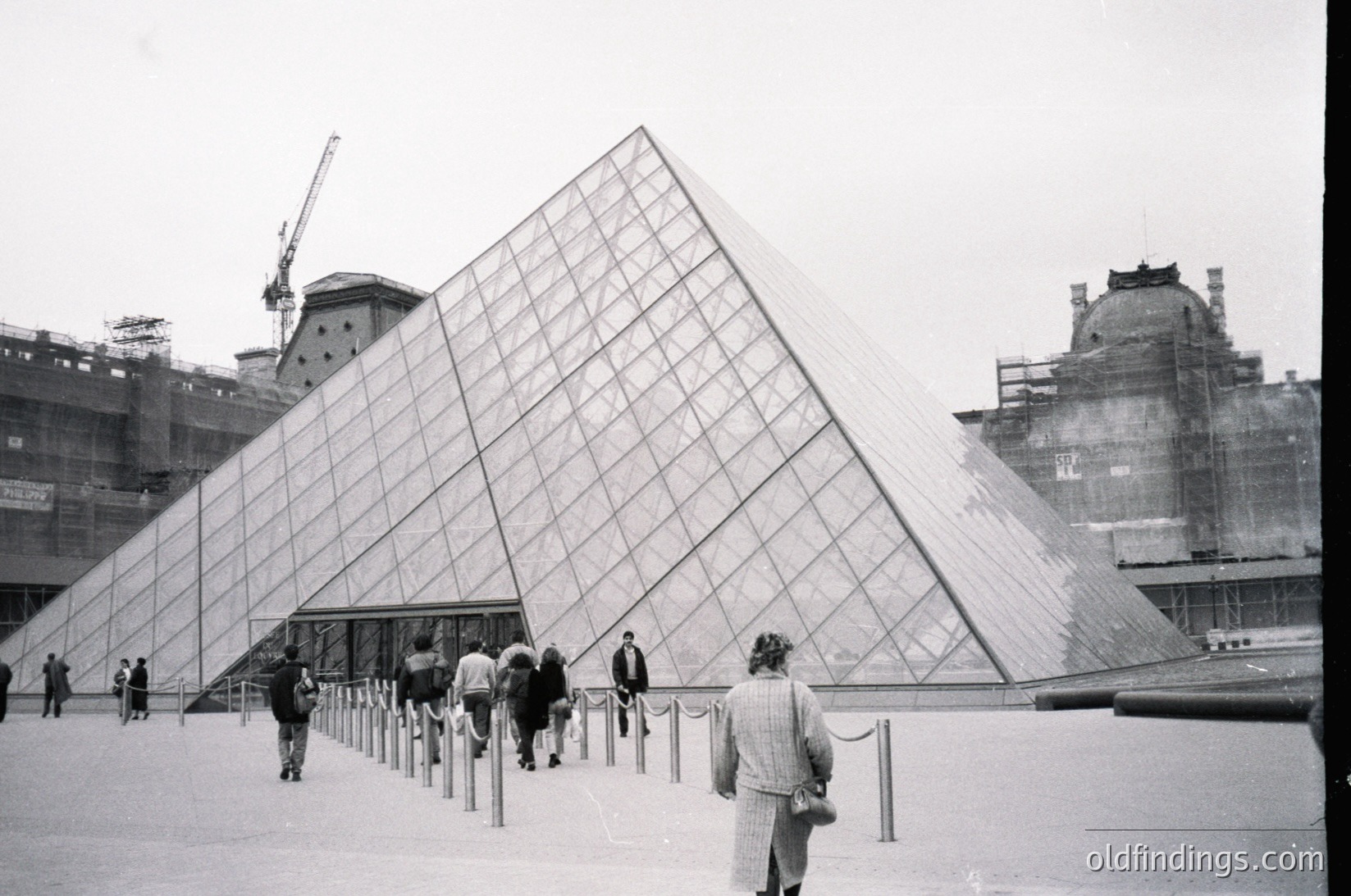 Iconic glass pyramid entrance of the **Louvre Museum**, Paris, France. Constructed by I.M. Pei in 1989, blending modernist design with historic architecture. Visitors explore the courtyard amid scaffolding and classical Louvre buildings.