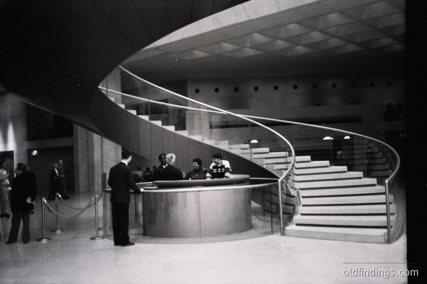 Mid-century modern reception desk with curved metal counter and sweeping staircase in a grand public building. Formal attire suggests official or institutional setting, likely a government or cultural hub. Architectural details hint at Brutalist or International Style, circa 1960s–1970s.
