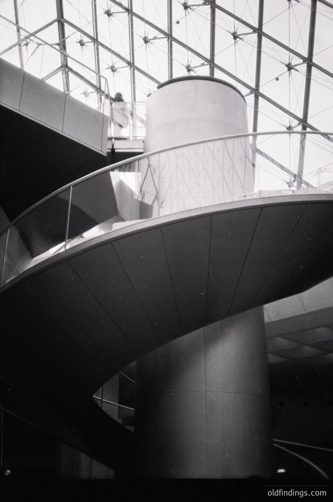 Modernist architectural interior featuring sweeping curved concrete ramps and cylindrical columns. Glass ceiling with structural grid allows natural light. Minimalist design with clean lines, likely a public or cultural space.