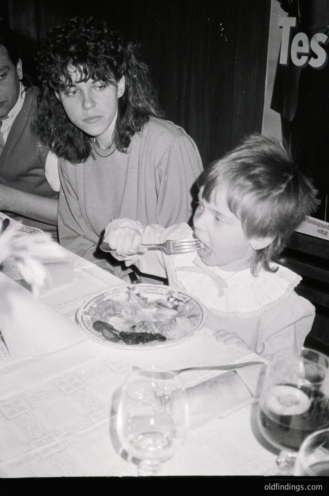 A woman and child seated at a formal meal, likely 1980s–1990s Western setting. The woman wears a high-neck blouse, while the child feeds himself with a fork and knife. Empty wine glasses and a partially eaten plate of food with meat and vegetables are visible. Background suggests an indoor dining event, possibly corporate or social.
