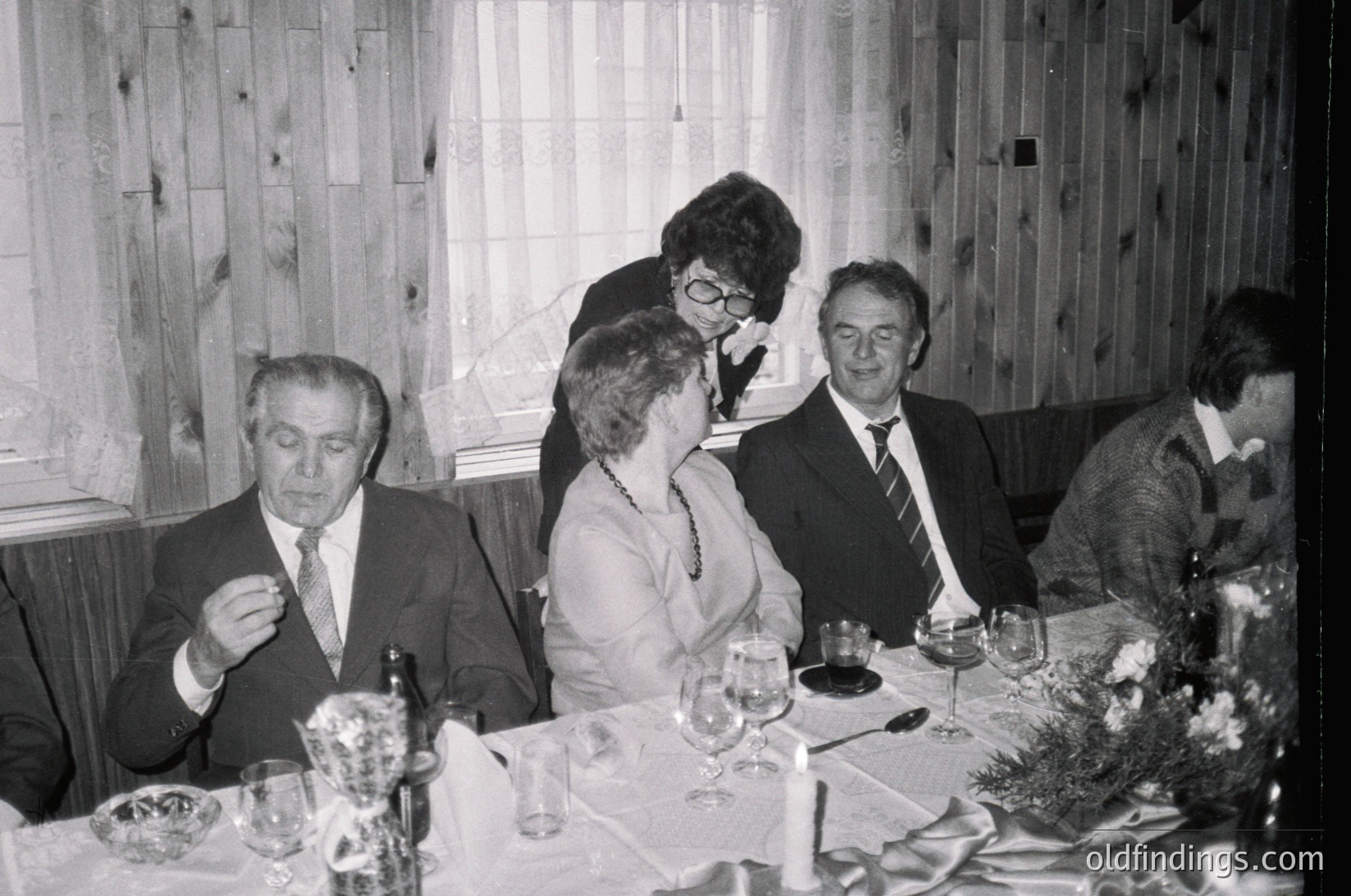 Mid-20th century formal gathering in a rustic wooden-paneled dining room. Six adults seated around a long table adorned with wine glasses, candles, and floral centerpieces. Men in suits, women in dresses; one woman leans forward in conversation. Soft focus suggests indoor lighting. Likely