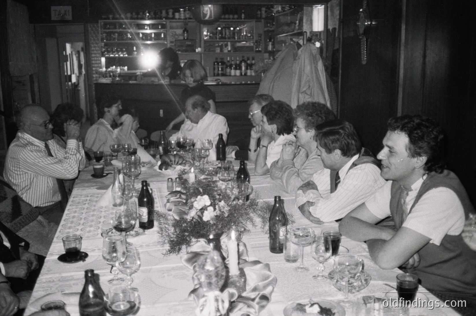 Black-and-white group dinner in a dimly lit, rustic indoor setting—likely a 1970s European gathering. Long table laden with wine bottles, glasses, and a floral centerpiece. Attendees in casual 70s attire (vests, button-ups) engage in conversation. Bar shelves with bottles in background suggest a social or celebratory occasion.