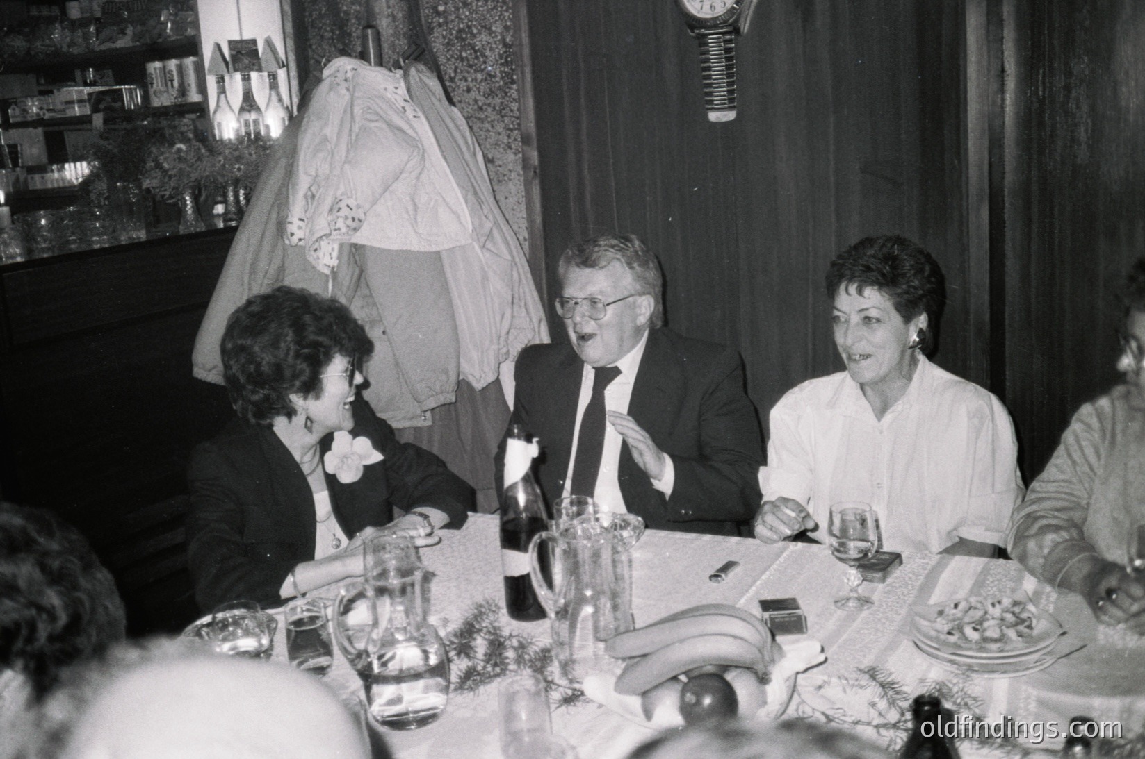 Black-and-white wedding reception scene featuring three central guests: a man in a suit laughing, flanked by women in formal attire. Veiled bride’s dress hangs above, suggesting recent ceremony. Table setting includes wine glasses, plates, and a bottle. Formal 1970s–1980s attire and indoor lighting indicate a mid-century European wedding. Ideal for vintage wedding archives or historical fashion research.