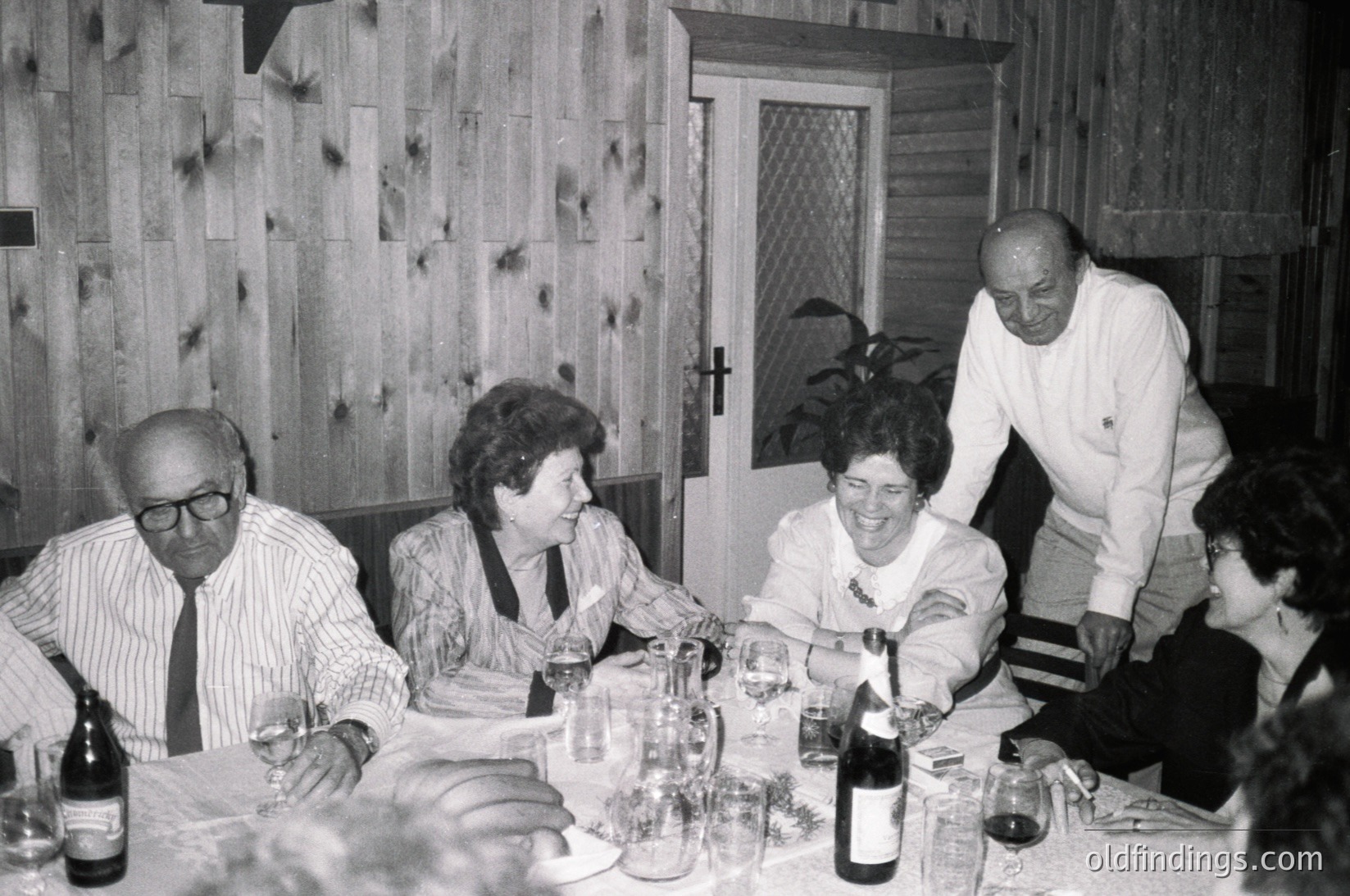 Mid-century indoor gathering at a wooden-paneled dining table, featuring five adults seated/standing around a wine-laden table. Men wear striped dress shirts; women in blouses with floral patterns. Bottles of wine, glasses, and a potted plant add context. Likely 1960s–1970s Western Europe or North America.