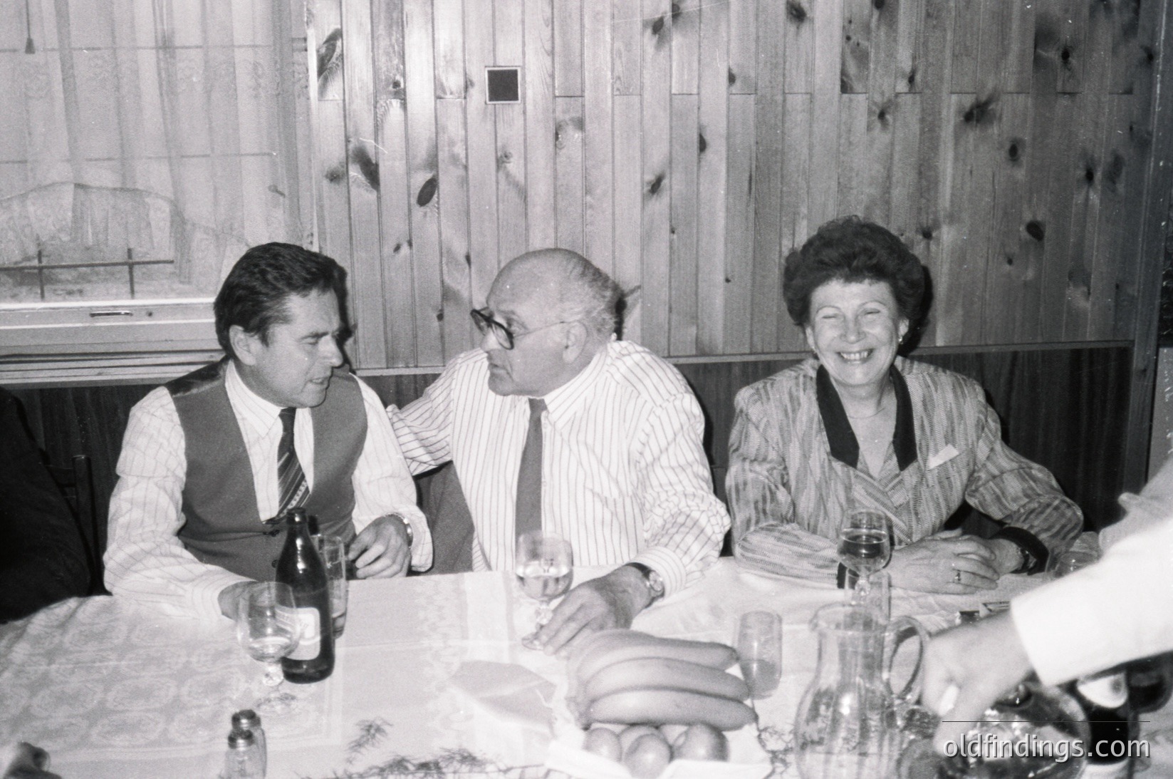 Black-and-white photo of three adults seated at a formal dining table, likely from the **1970s**. Wood-paneled walls and vintage tableware suggest a mid-century European setting. The man in the center wears round glasses and a striped shirt, while the woman on the right smiles warmly. Glasses of wine and a bottle of champagne indicate a celebratory occasion.