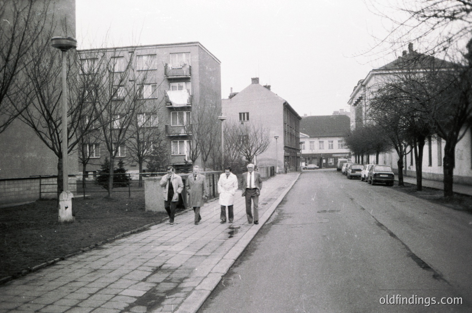 Three men in winter coats walk along a paved sidewalk beside a residential street lined with mid-century concrete apartment blocks and parked cars. Leafless trees and overcast skies suggest late autumn/winter.