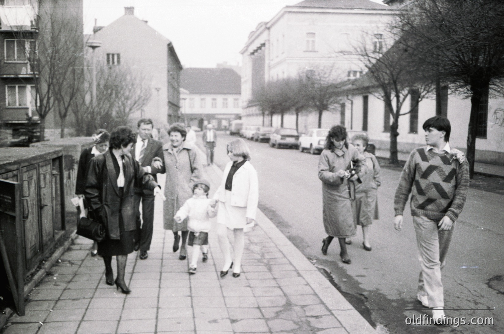 A mid-20th century street scene featuring a diverse group of people walking on a paved path lined with leafless trees. Adults in coats and hats, children in light jackets, and a man in a patterned sweater. Soviet-era urban architecture with multi-story buildings in the background. Likely Eastern Europe, 1960s-1970s.