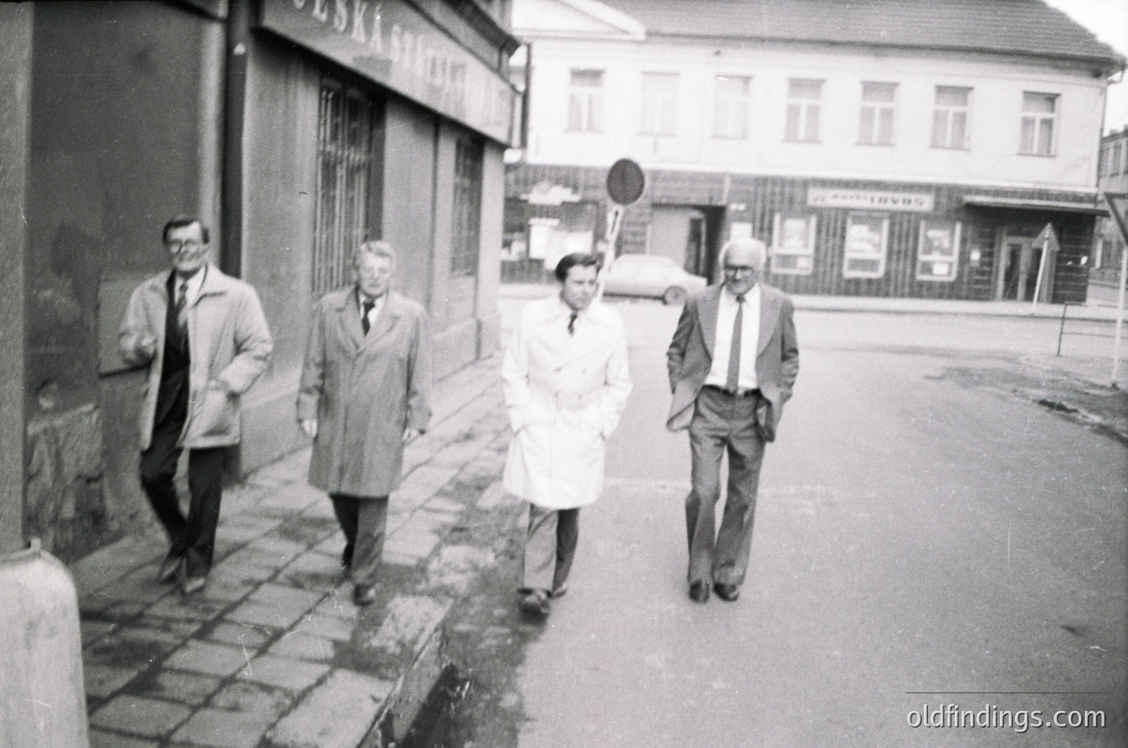 Four individuals in 1960s-era attire—men in coats and ties, one woman in a white coat—walk along a wet urban sidewalk. Soviet-style architecture with closed shops and a "1900" signage detail visible. Likely Eastern Bloc city, mid-20th century.