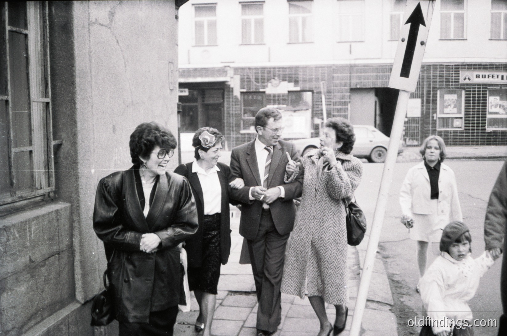 Mid-20th century street scene featuring a group of adults and a child in formal attire, likely 1960s–1970s. Men and women in suits, coats, and hats gather near a building with a visible "Hof" sign. A large arrow sign in the foreground suggests directional guidance. Urban setting with classic cars and storefronts.