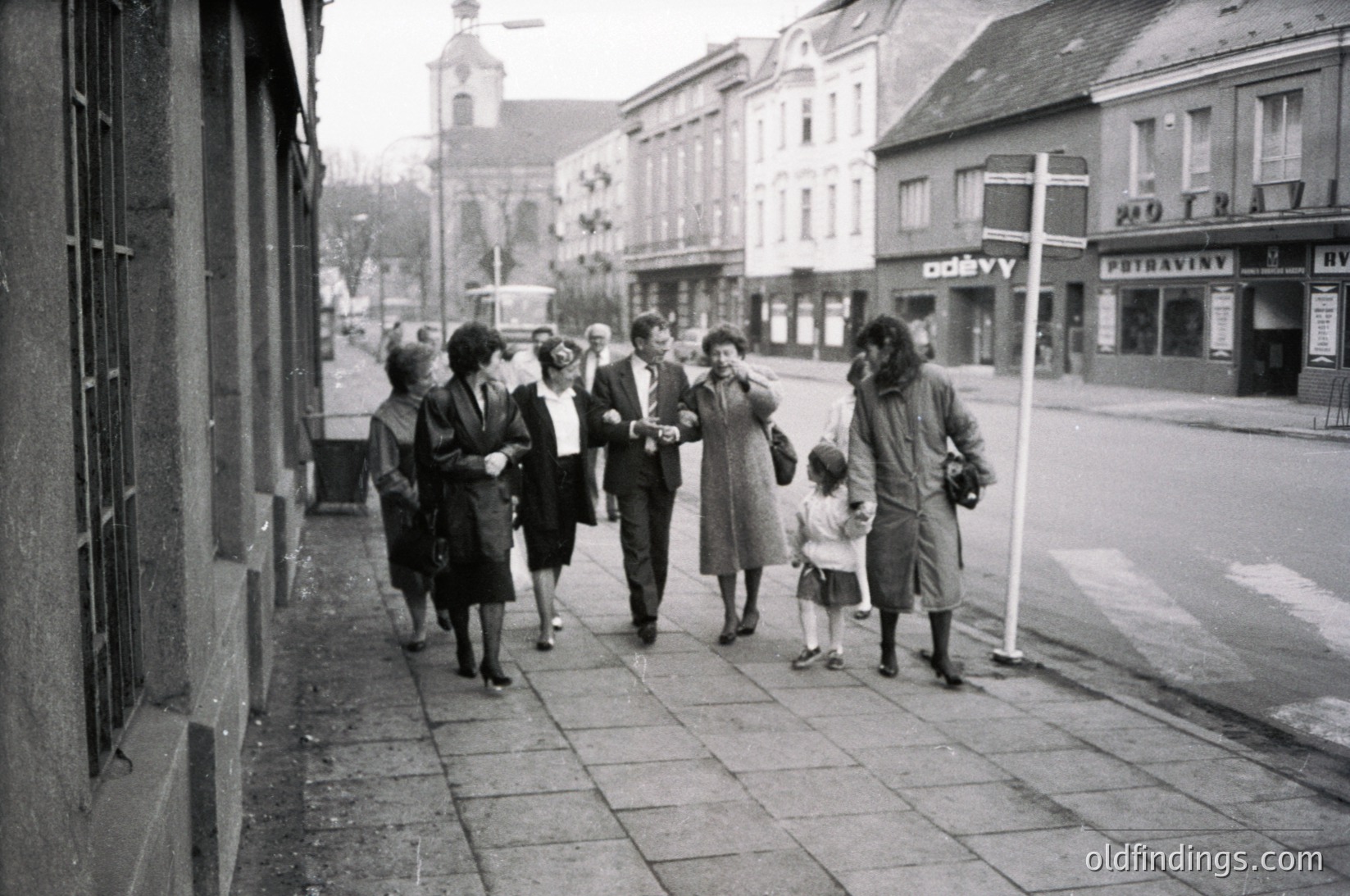 Mid-20th century street scene with group of adults and child walking on paved sidewalk. Architectural details include closed shopfronts with signs in Cyrillic script (, ). Uniform clothing suggests post-war era, likely 1950s-60s. Urban setting with visible tram tracks and vintage signage (, ).