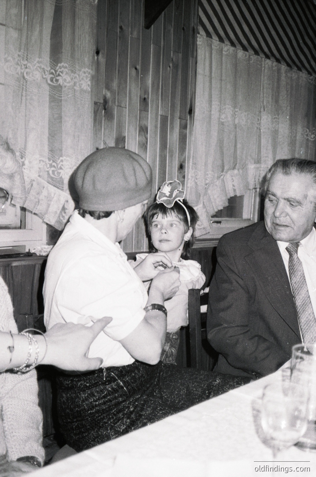 Black-and-white indoor scene from the 1960s–70s featuring a man in a suit adjusting a child’s hat, likely at a formal event. Wood-paneled walls and patterned wallpaper suggest mid-century European decor. Glassware and table settings indicate a reception or gathering.