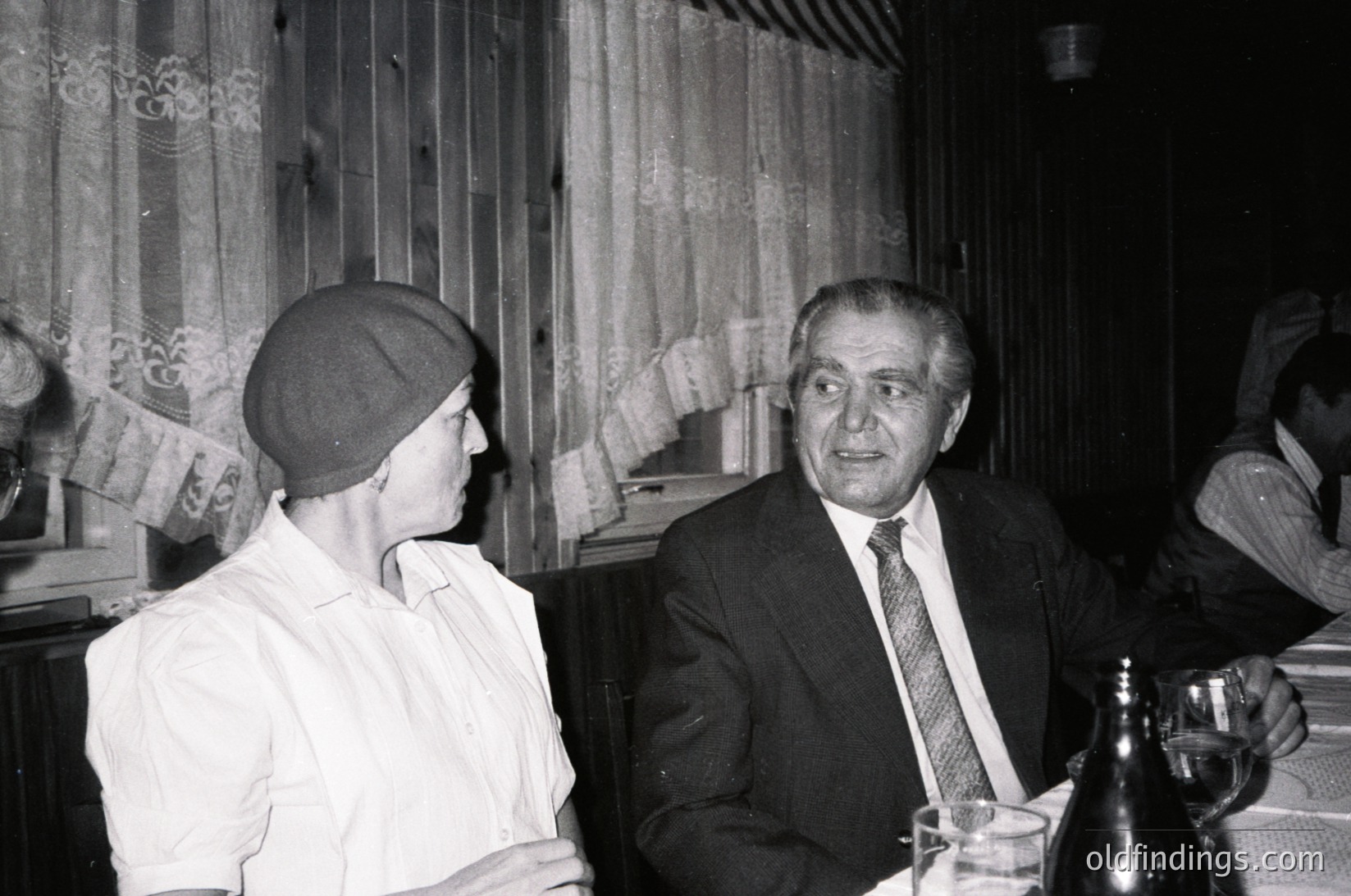 Two individuals in mid-20th-century formal attire converse indoors, likely at a social gathering. The man wears a suit, tie, and cap; the woman sports a cap and blouse. Wood-paneled walls and a patterned curtain suggest a classic European café or restaurant setting. Bottle and glass on the table indicate a drink. é