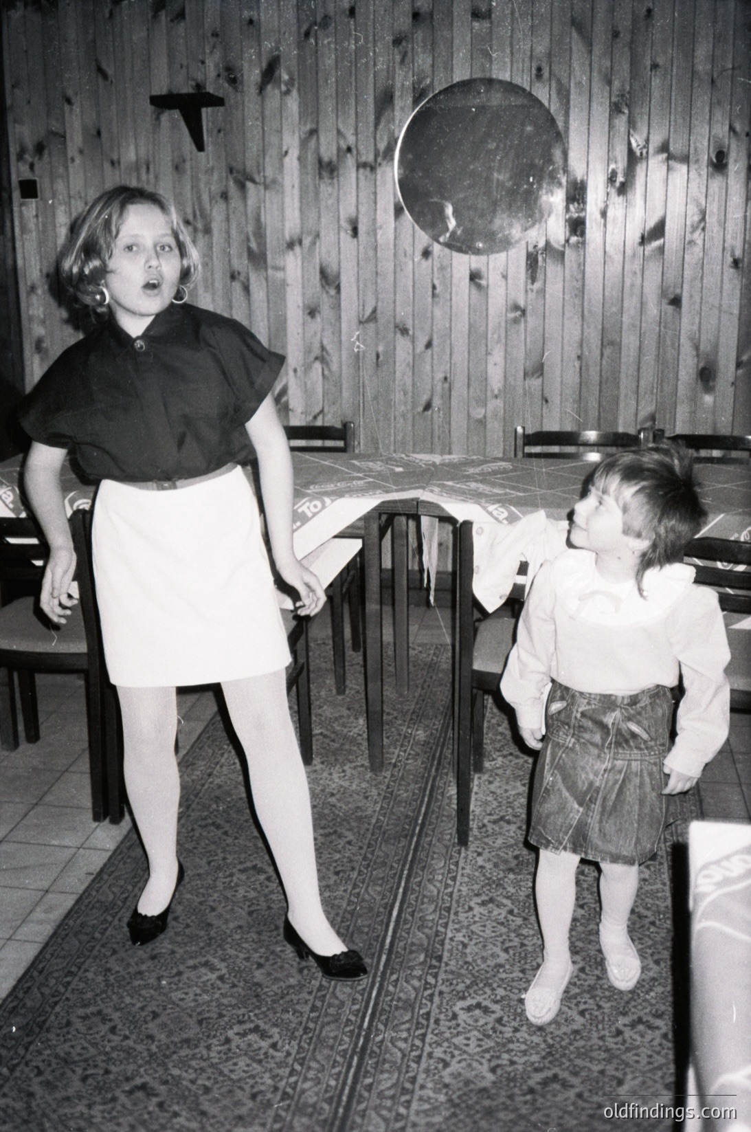 Two children in a wooden-paneled indoor setting, likely a 1960s-70s dining area. The girl in a black collar dress and white skirt stands near a table, while the boy in a striped shirt and shorts reaches upward. Circular wall clock and patterned tablecloth visible.