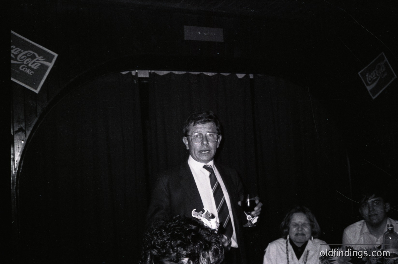 Man in formal attire speaking at a podium under stage lighting, flanked by floral arrangements. Coca-Cola signage visible in background, suggesting 1970s–1980s event setting. Indoor venue with dim lighting and partial audience in view.