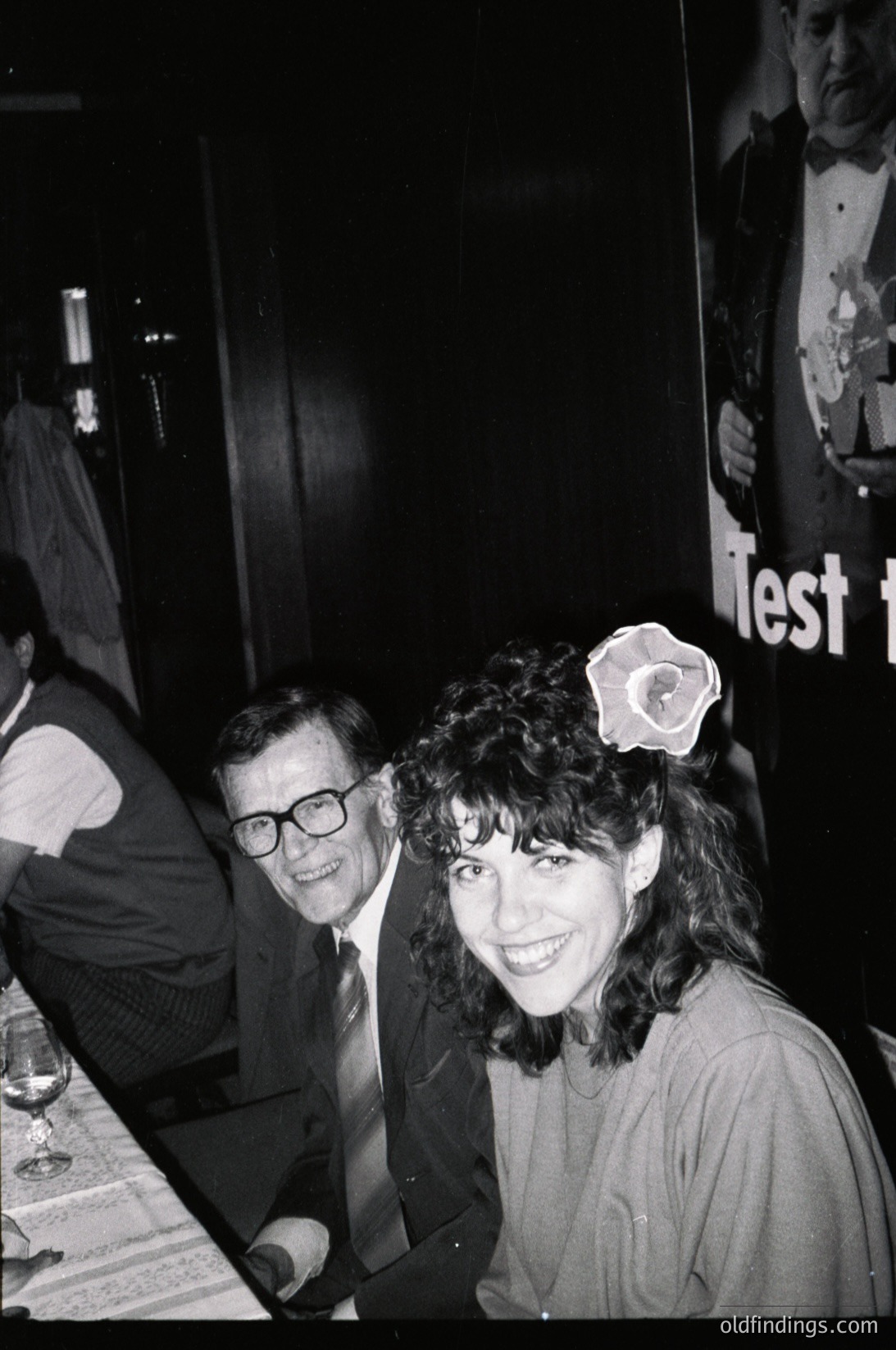 Black-and-white candid shot of two people at an indoor event, likely a 1970s–1980s social gathering. The woman wears a flower hair accessory and a layered top, while the man in glasses sports a suit and tie. Posters in the background suggest a theatrical or concert venue. Candid expressions and lighting evoke a lively atmosphere.
