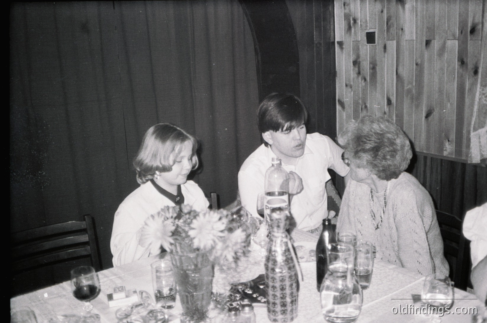 Three individuals seated at a table in a mid-century indoor setting, likely a 1960s-1970s home or gathering. The woman on the right wears a patterned blouse, the man in the center a collared shirt, and the child on the left a simple dress. Decorative glass bottles and a vase with dried flowers adorn the table. Wooden paneling and a curved arch frame the background.