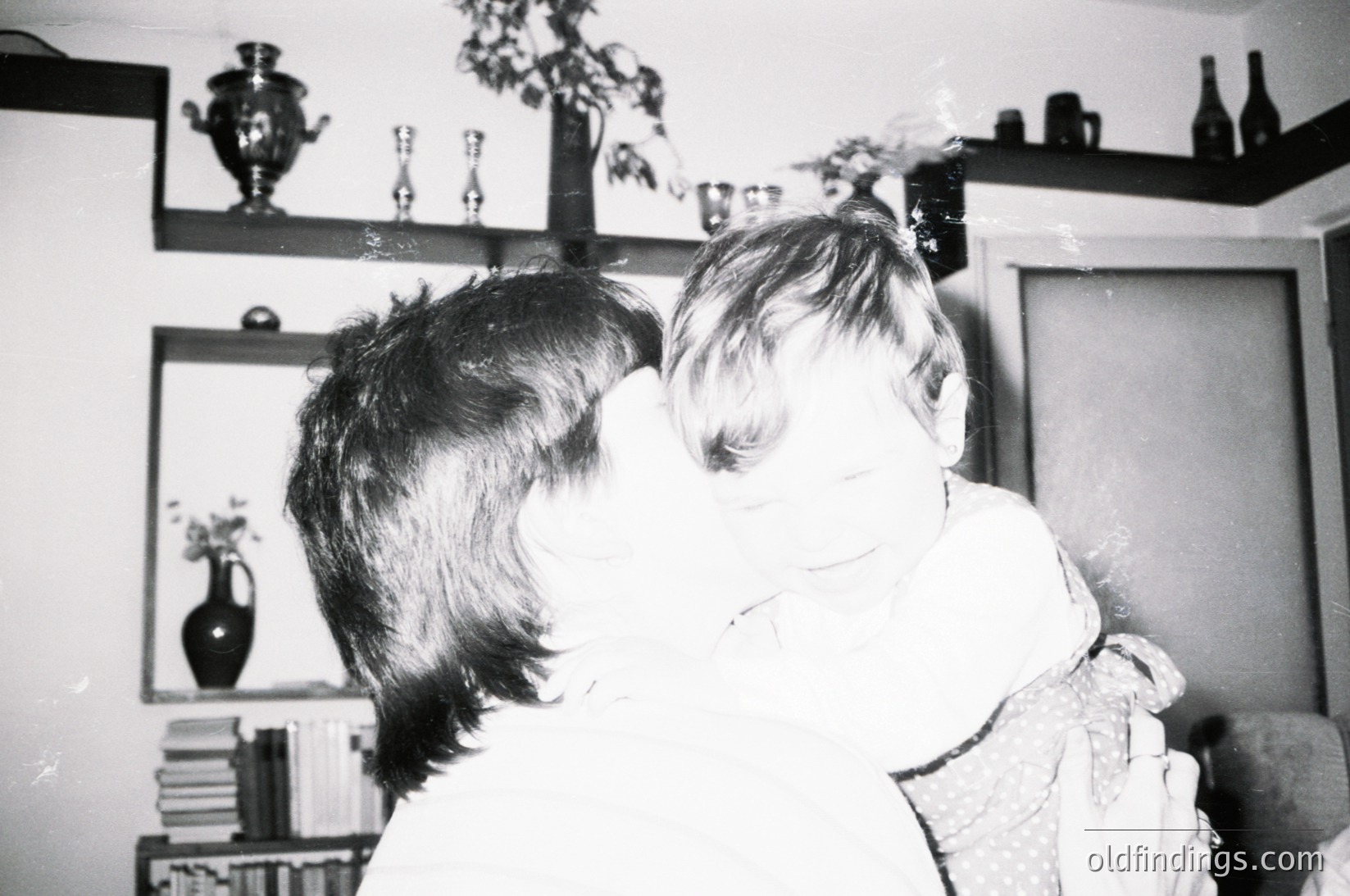 A tender black-and-white moment of a child hugging a large, shaggy dog indoors. Shelves behind display vintage glassware, books, and decorative items, suggesting a mid-20th-century home. Warm, intimate family scene evokes nostalgia.