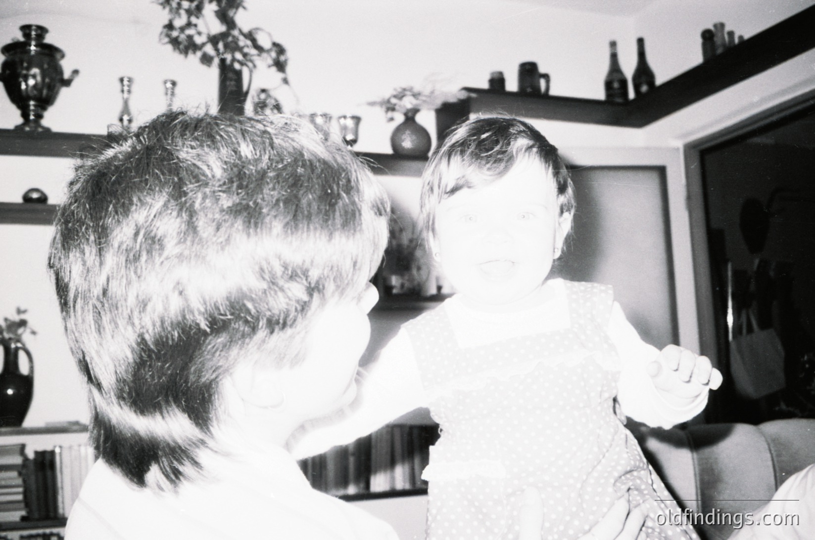Vintage black-and-white interior shot of two people embracing in a living room, likely 1960s–1970s. Decor includes ceramic vases, framed photos, and a shelf with bottles. Warm, intimate family moment captured in mid-embrace.