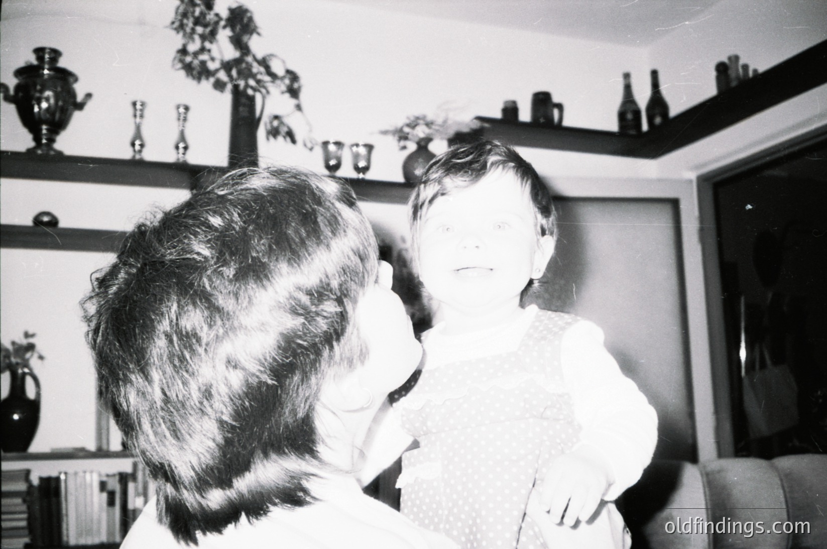 Mid-20th century indoor portrait: an adult and child share a tender moment in a cluttered living room. Shelves display glassware, bottles, and floral arrangements; a sofa and armchair frame the scene. Likely
