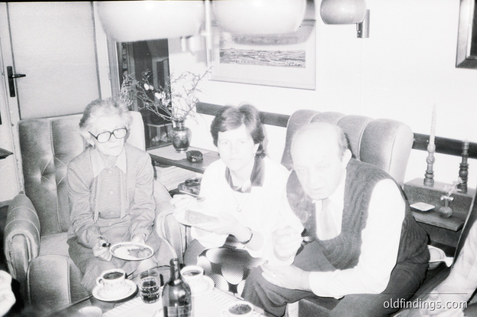 Three adults seated indoors in a mid-century living/dining area, likely 1960s–1970s. Glasses, plates, and a wine bottle suggest a casual meal. Soft lighting, floral centerpiece, and framed artwork hint at mid-century design.