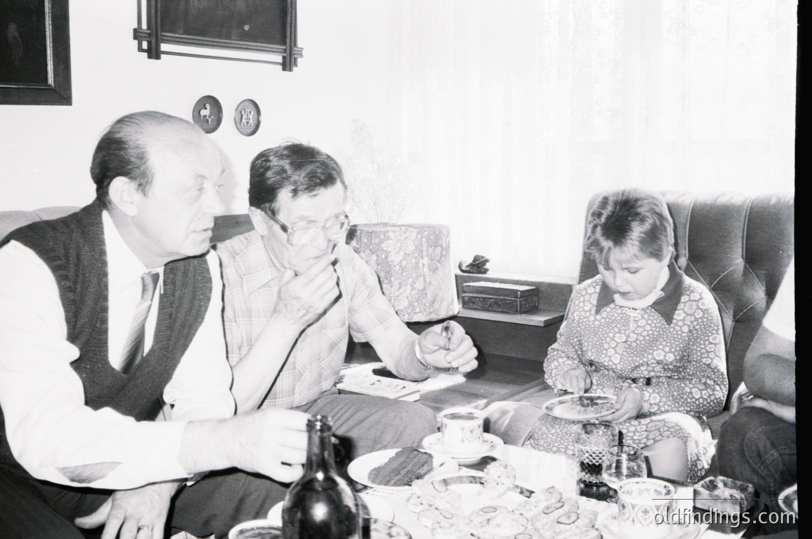 Family gathering in a mid-century living room, featuring three adults and a child seated around a low coffee table. The adults wear patterned sweaters and formal attire, while the child wears a floral dress. A wine bottle, glasses, and scattered coins on the table suggest a casual yet intimate occasion. Decor includes a framed clock and patterned wallpaper, indicative of 1960s–1970s domestic life.