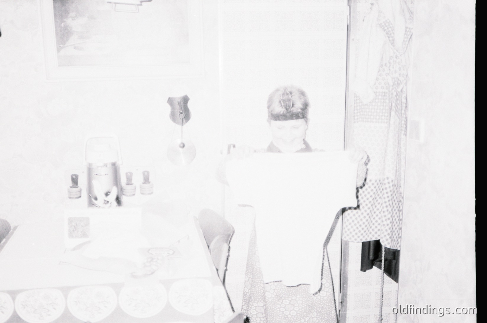 Vintage black-and-white interior shot of a minimalist 1960s–70s living space. Clean lines, geometric furniture, and a single framed abstract art piece on a stark white wall. Mid-century modern decor includes a cylindrical table lamp, geometric cushions, and a blurred figure in a patterned dress.
