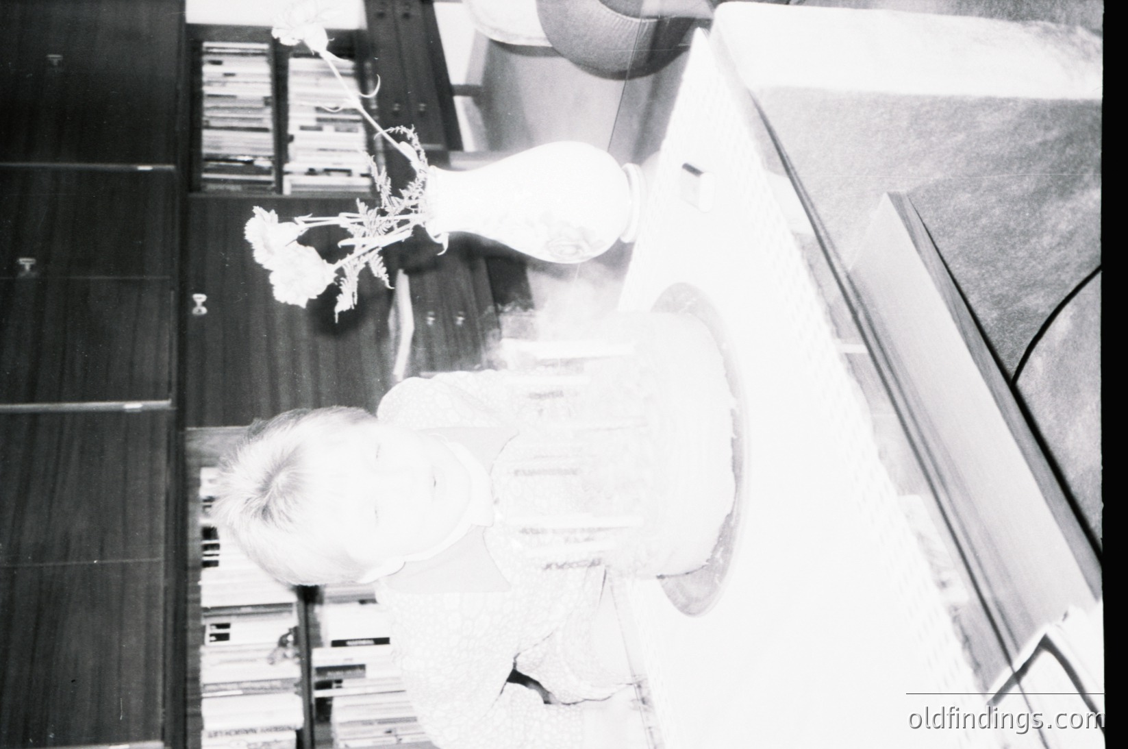 Vintage black-and-white shot of a child sitting on a wooden floor, surrounded by stacked bookshelves. A vase with a single white flower rests on a low table beside them, creating a serene indoor study scene. Likely mid-20th century, evoking midcentury design aesthetics.