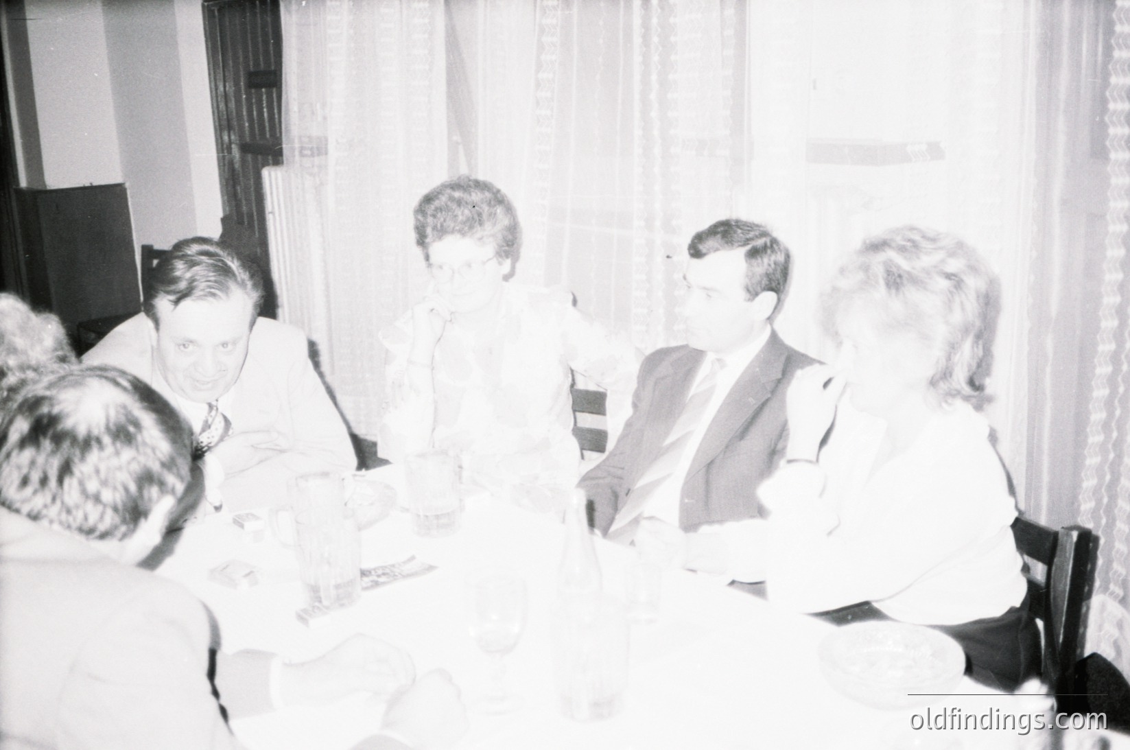 Five adults seated around a table in a dimly lit indoor setting, likely a formal gathering or meeting. Men wear suits, women in blouses/dresses. Table holds papers, glasses, and a microphone, suggesting a discussion or speech. Curtains and framed art hint at a conference room or event hall. Mid-20th century (1950s–1970s) style.