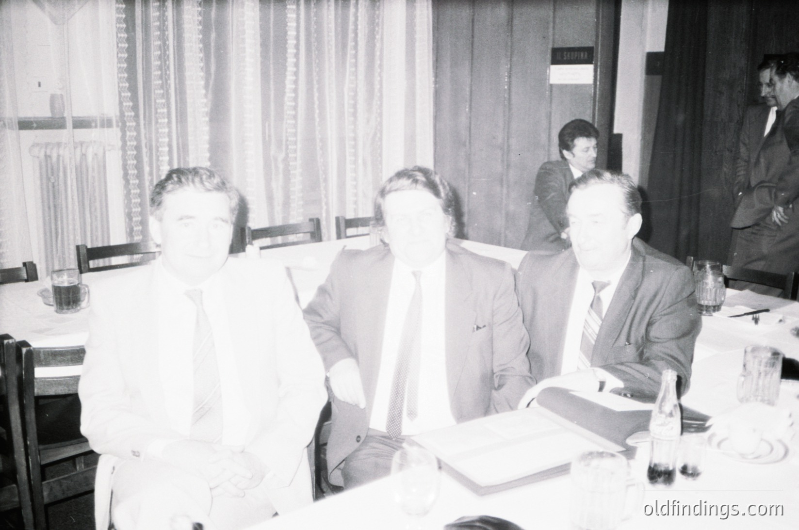 Three men in formal attire—dark suits, ties, and dress shirts—engage in discussion at a rectangular table in an indoor setting, likely a conference or meeting room. The table holds documents, pens, and ashtrays. Curtains with a subtle pattern frame the windows behind them. Style and lighting suggest a mid-20th-century office environment, possibly 1950s–1970s.