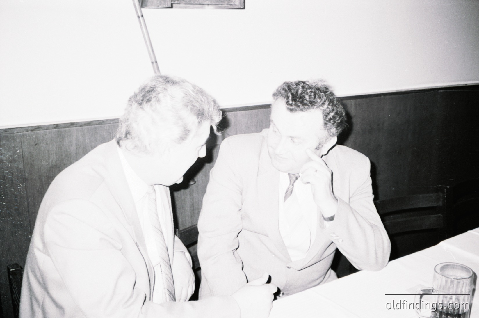 Two men in formal attire—white button-down shirts, ties, and blazers—engage in conversation indoors, likely mid-20th century ( ). One gestures with his hand, while the other leans in attentively. Plain wooden paneling and a glass ashtray on the table suggest a formal or semi-formal setting, possibly a conference or office.
