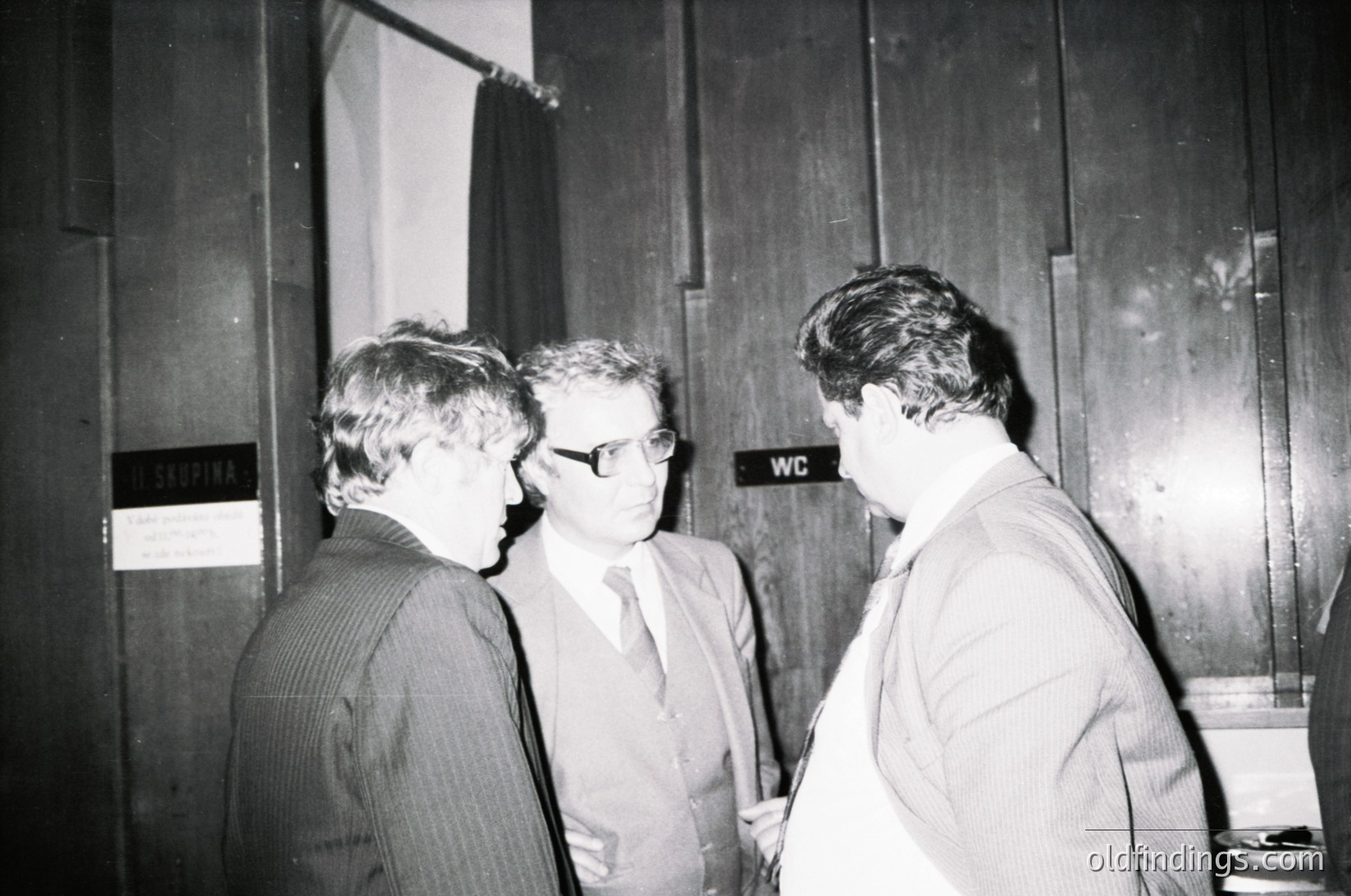 Three men in formal attire—striped suit, white shirt with tie, and light blazer—engage in conversation near a wooden-paneled wall with "WC" signage. Mid-20th century office or institutional setting, likely 1960s–1970s.