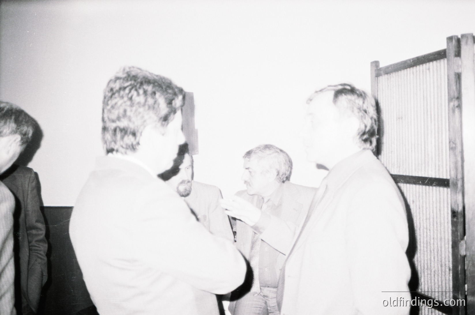 Black-and-white photo of a group of men in formal attire, likely 1960s–1970s, gathered around a projector screen. One man in a light-colored suit holds a cigarette, while others face the screen. Industrial setting with metal mesh wall and plain walls.